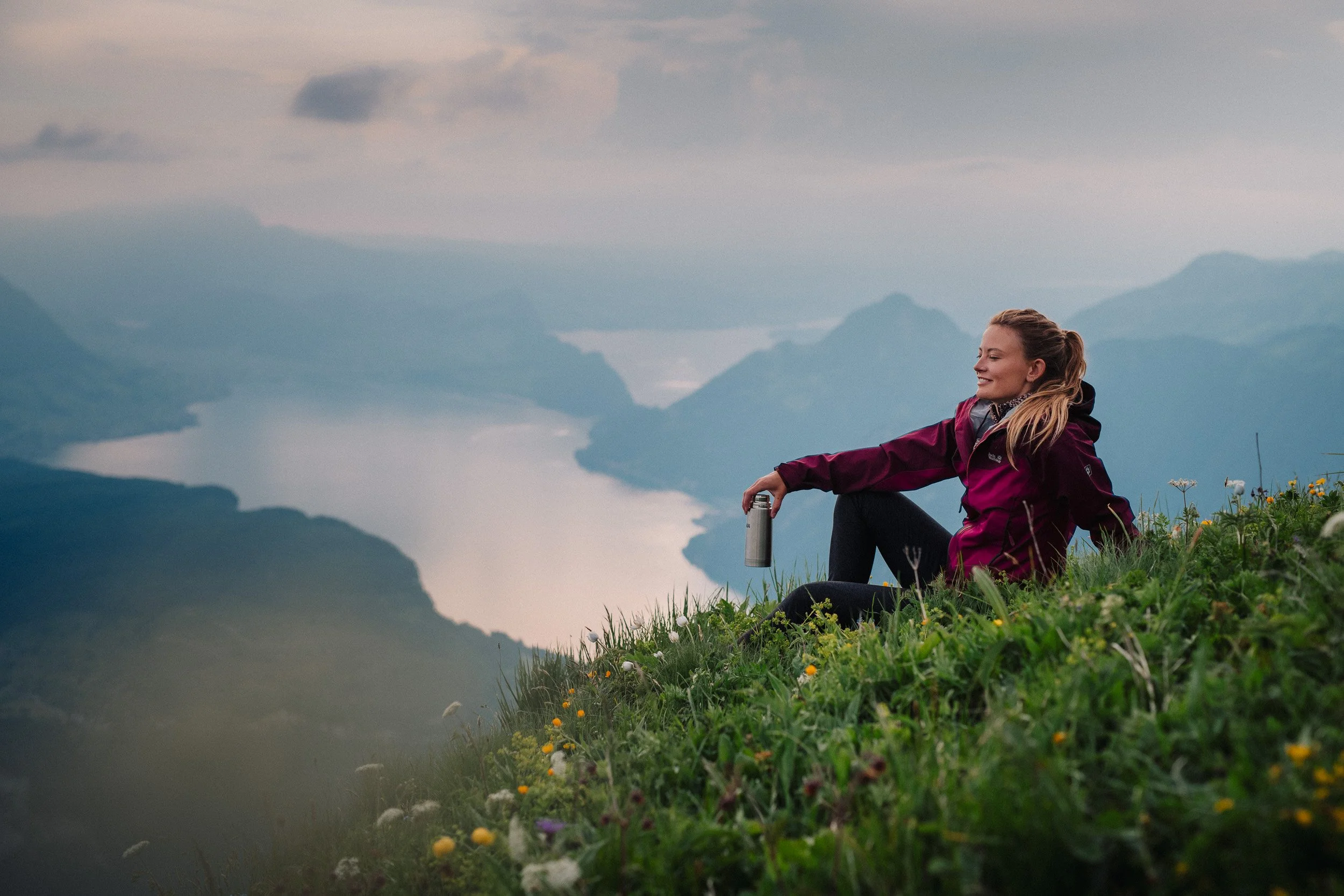 A woman sitting on a grassy hillside overlooking a mountain lake at sunrise, holding a thermos and smiling