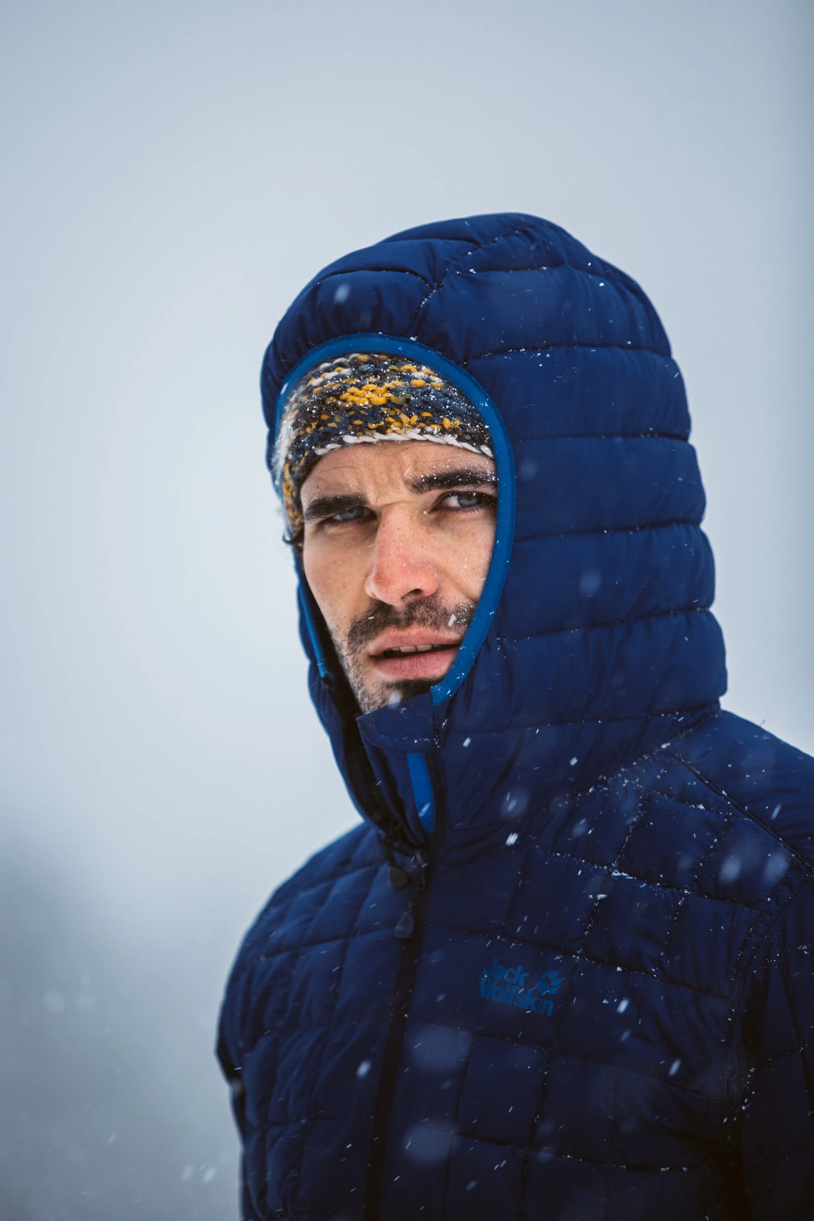 A man in a blue Hooded jacket and patterned knit cap looking into the camera with a serious expression on a snowy day.