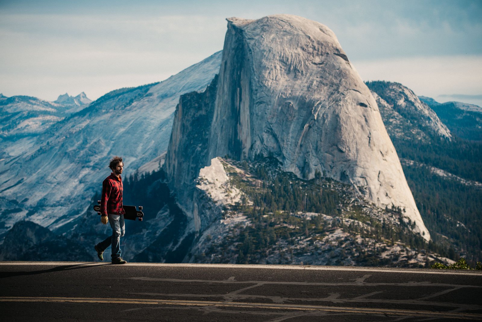 A person holding a skateboard walking along a scenic mountain road with a large granite rock formation in the background.