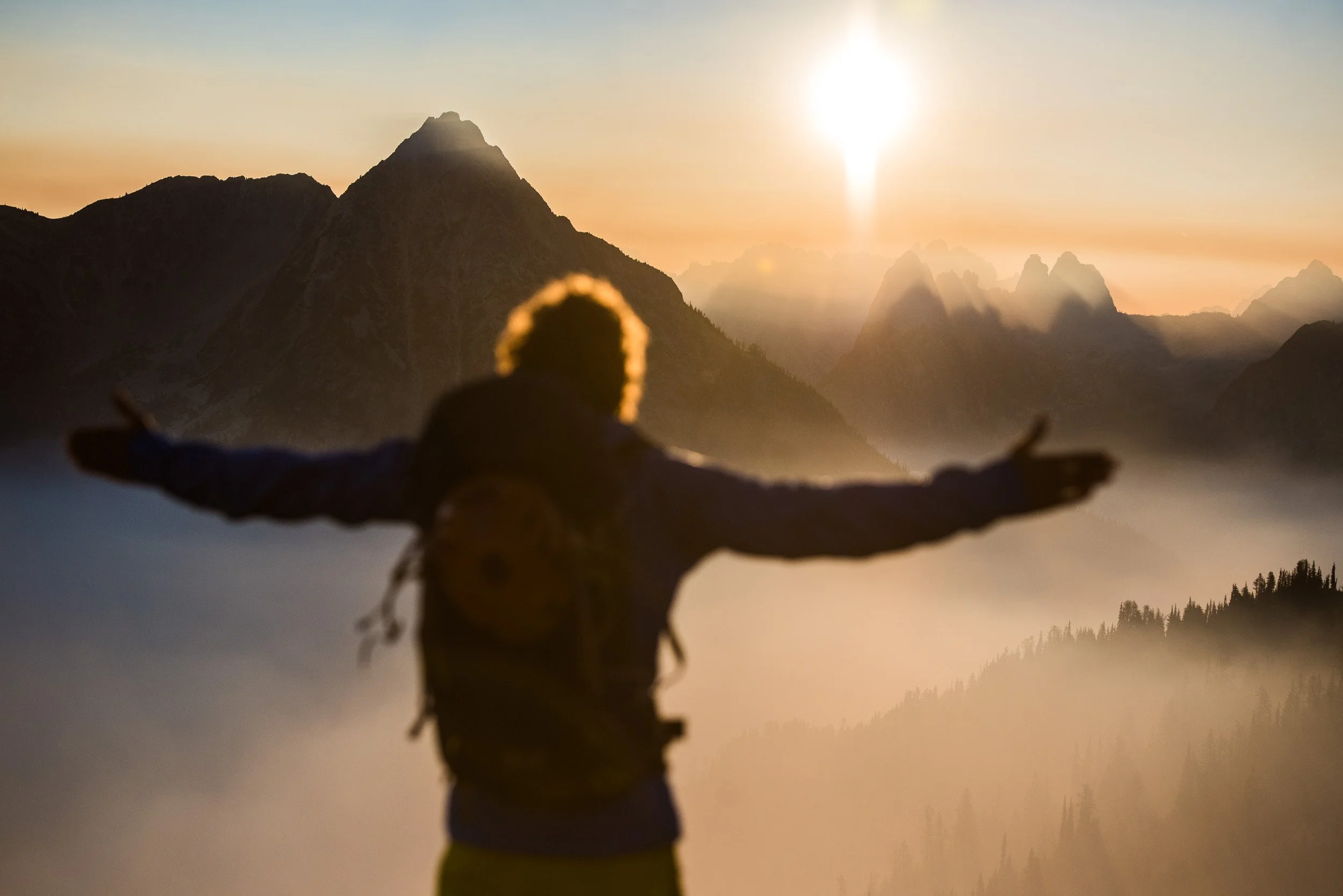Person standing with arms outstretched facing mountain landscape at sunset, with sun low in sky and misty mountains in background.