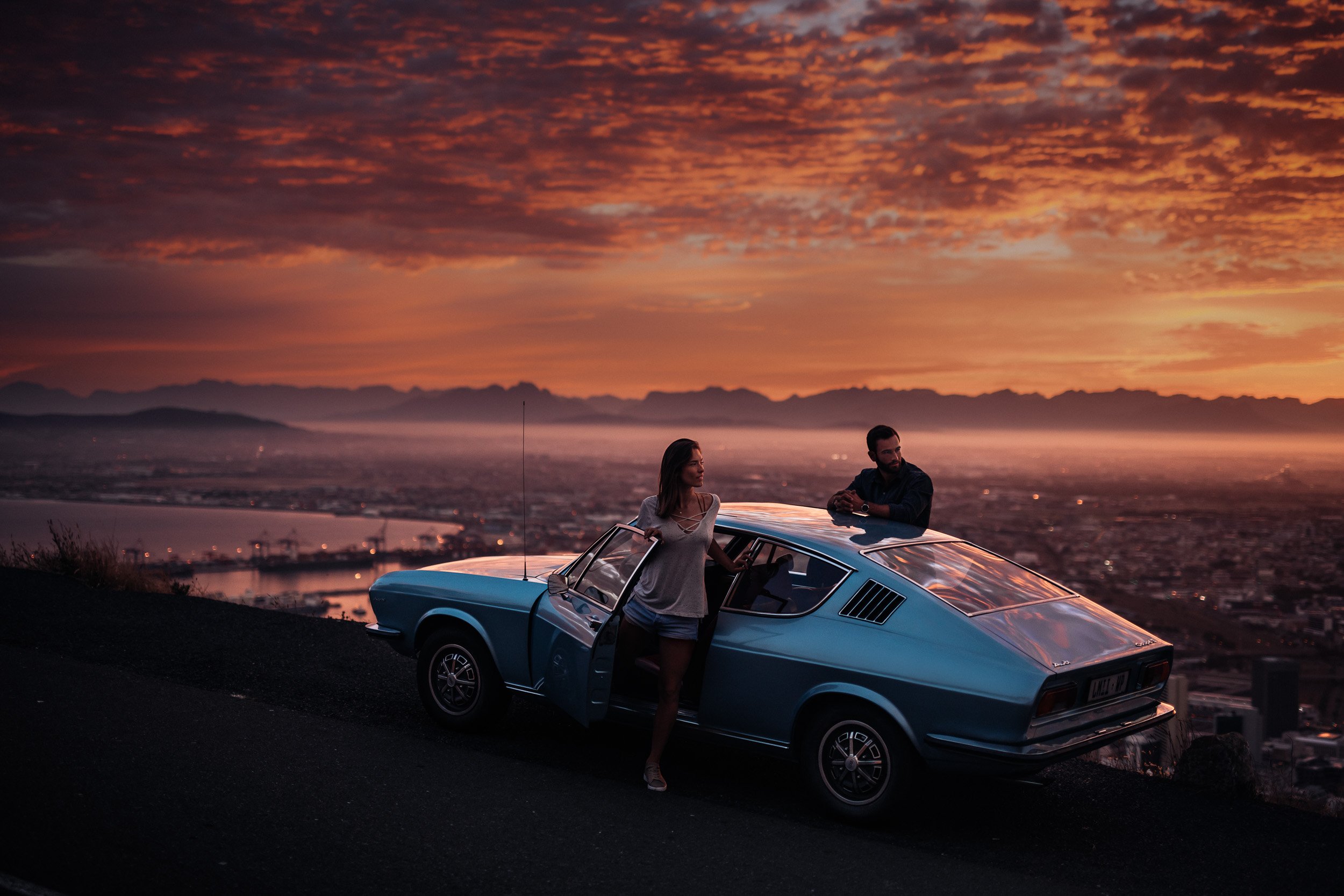 A vintage blue car parked on a hill at sunset with a woman standing outside and a man leaning on the car, overlooking a cityscape and distant mountains.