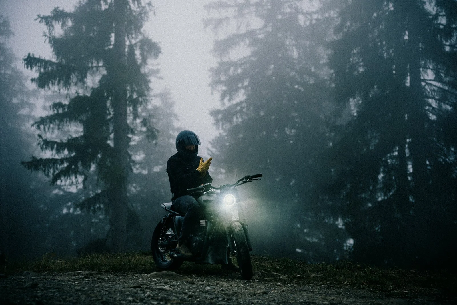 A person wearing a helmet and gloves sitting on an electric motorcycle in a foggy forest, checking their phone with the motorcycle's headlight on.