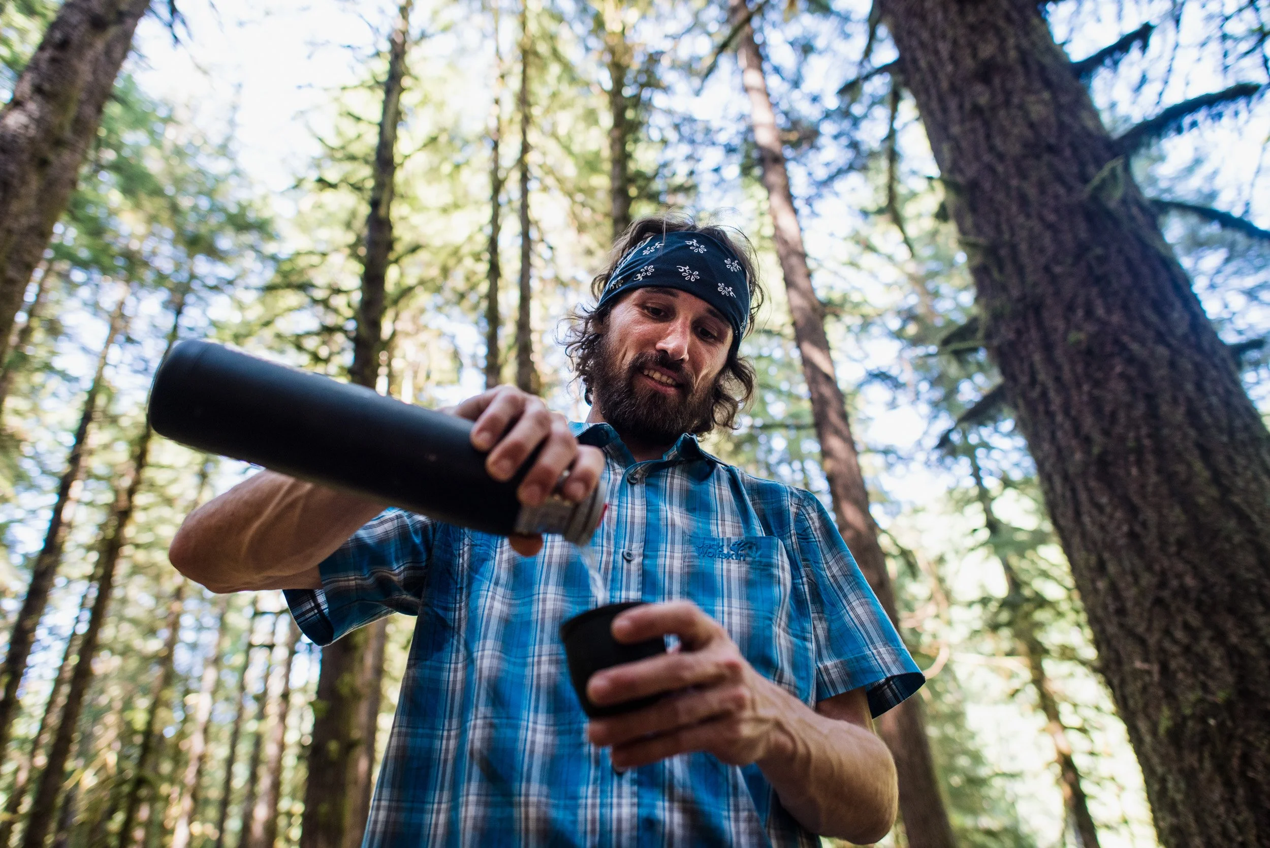 A man with a beard and long hair wearing a plaid shirt and bandana in a forest pouring a beverage from a thermos into a cup.