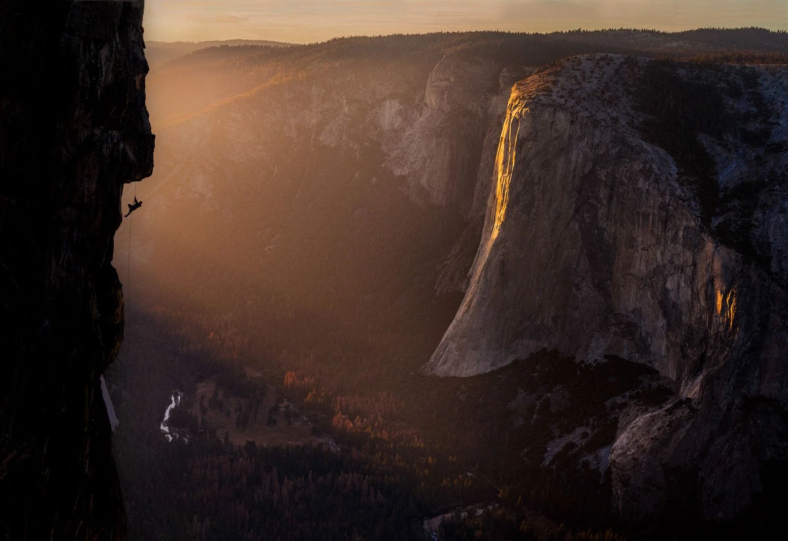 A rock climber ascending the dark face of a steep cliff during sunset, with a panoramic view of a canyon and forest below.