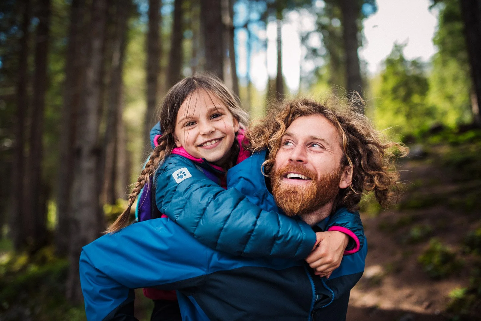 A man with curly hair and a beard carrying a young girl with braided hair on his shoulders in a forest