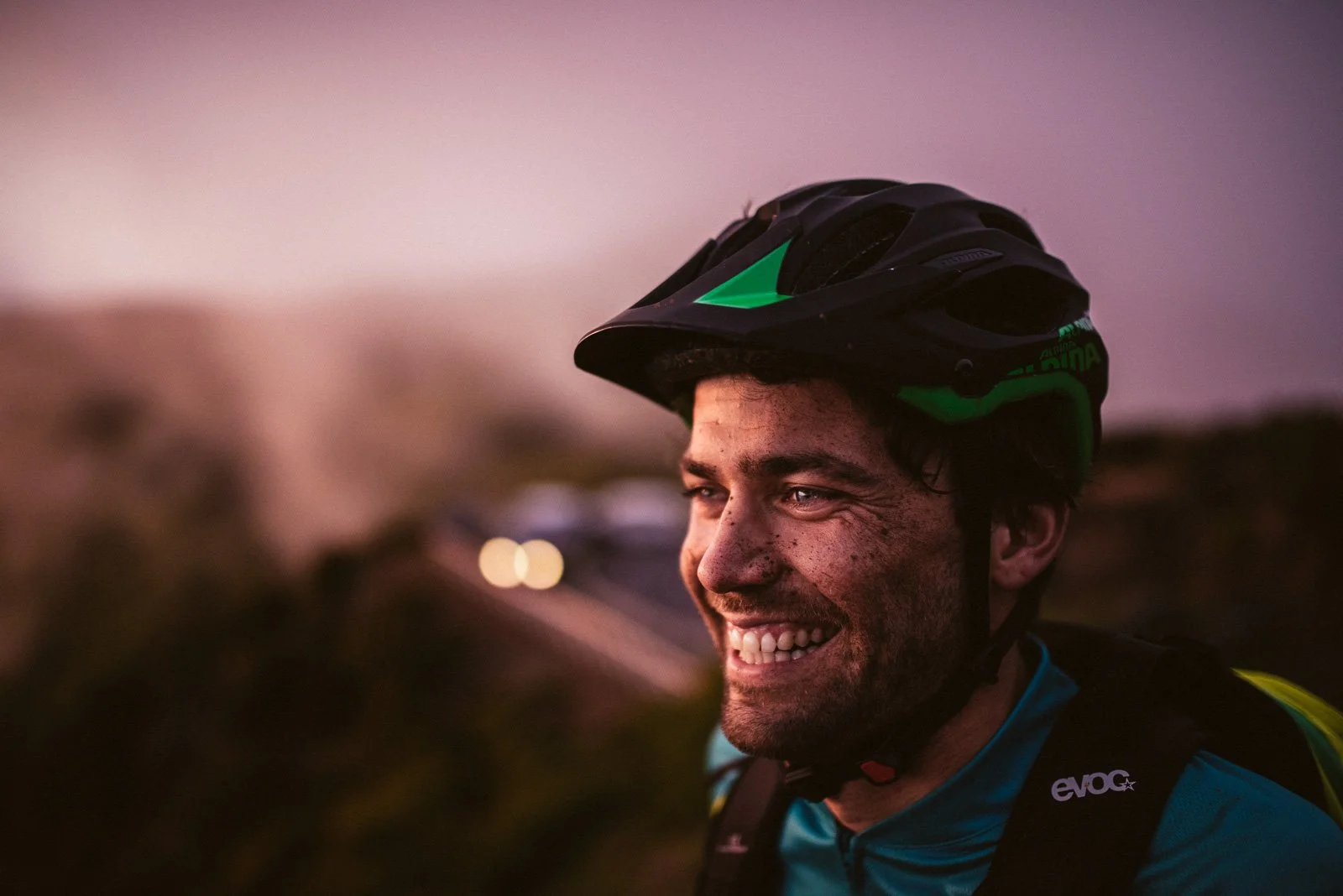 A smiling man wearing a black and green bicycle helmet outdoors at dusk.