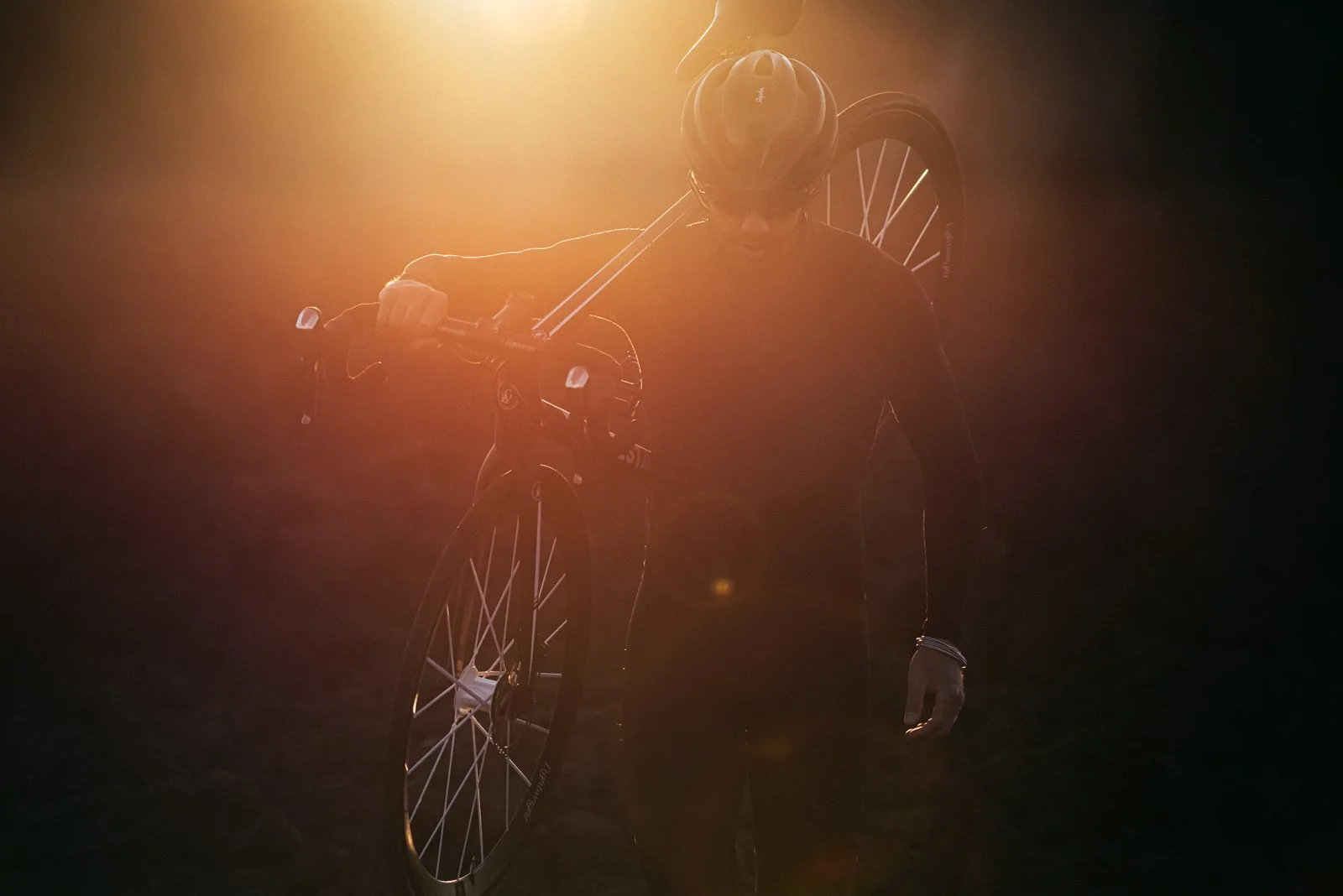 A person wearing a helmet carrying a bicycle on their shoulder, with the sun shining behind them, creating a backlit silhouette in a dark environment.