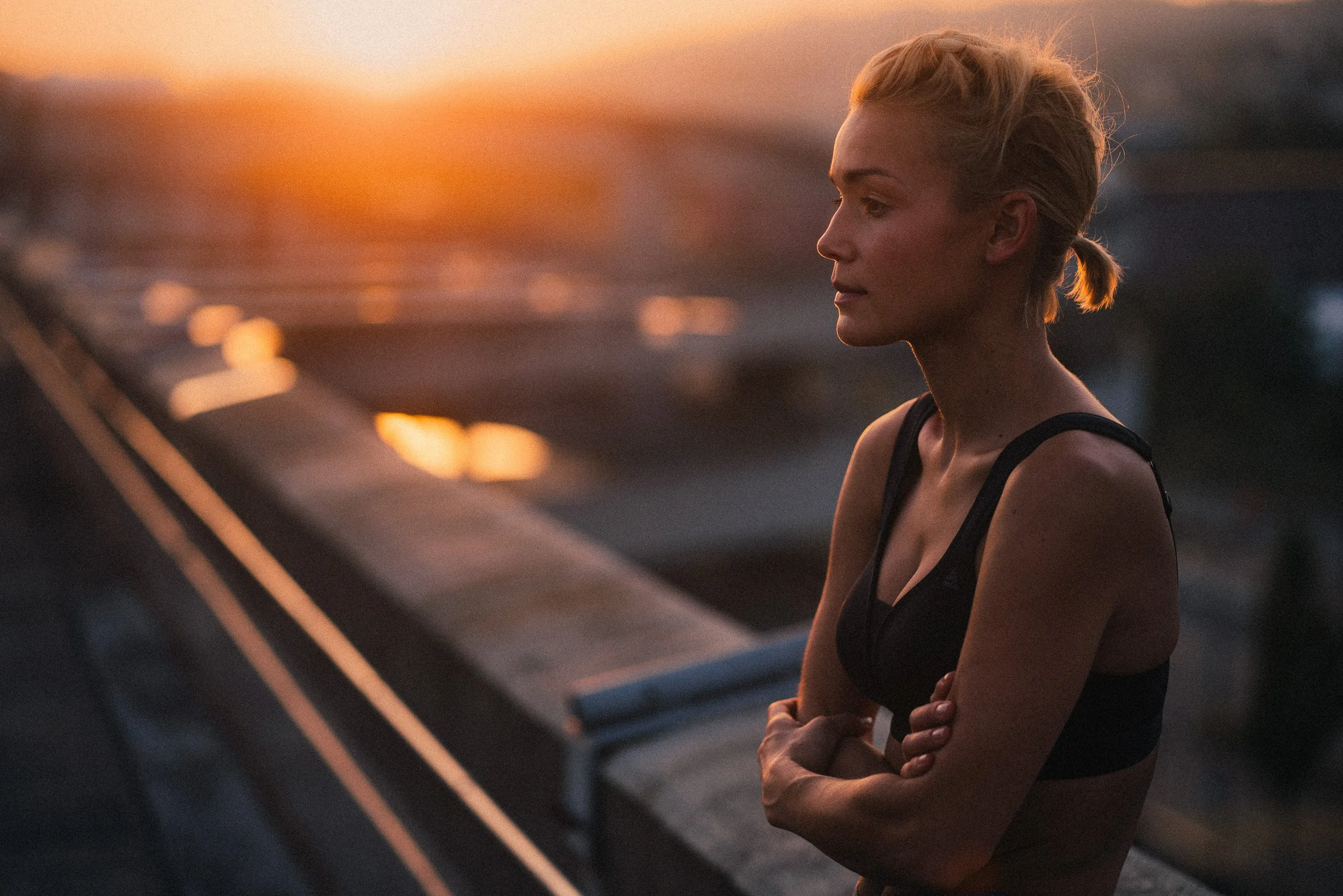 A woman in a black sports bra with blonde hair tied back, standing outdoors during sunset, with her arms crossed and looking contemplatively to the side.
