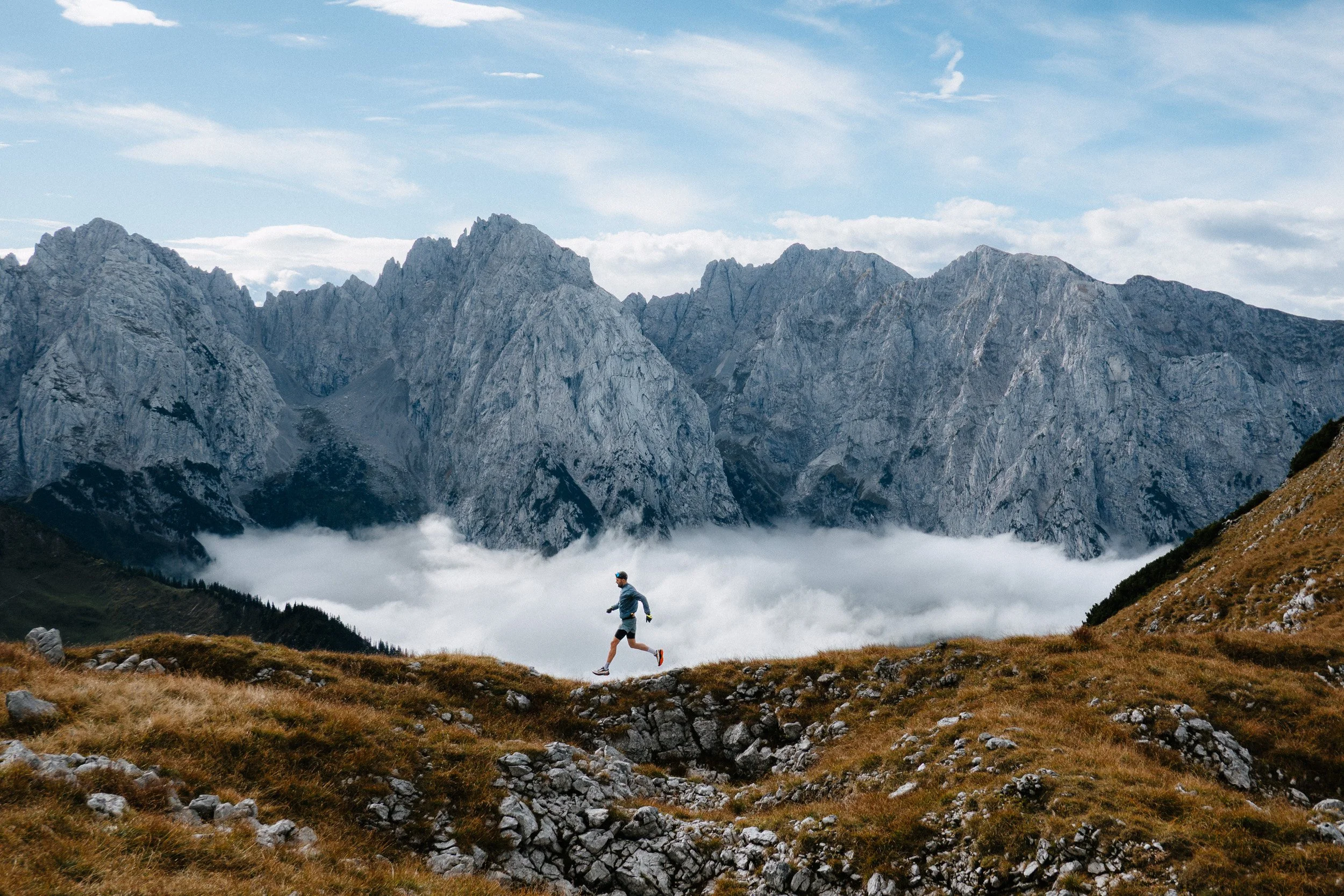 Person jogging on a trail in a mountainous landscape with rocky terrain, green patches, and clouds below distant rugged mountains under a partly cloudy sky.