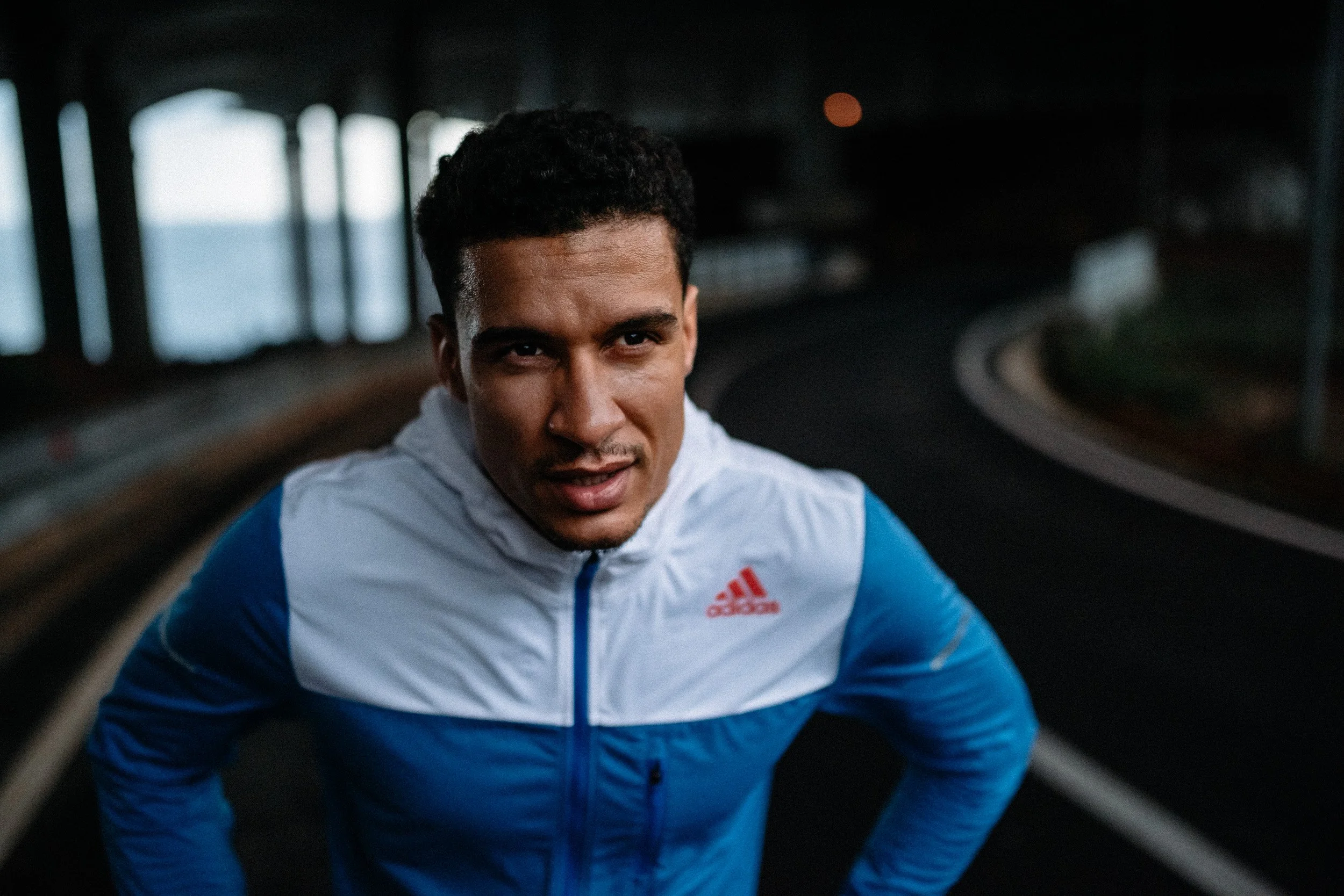 A young man in a blue and white Adidas sports jacket running on a dark, curved road under a bridge.