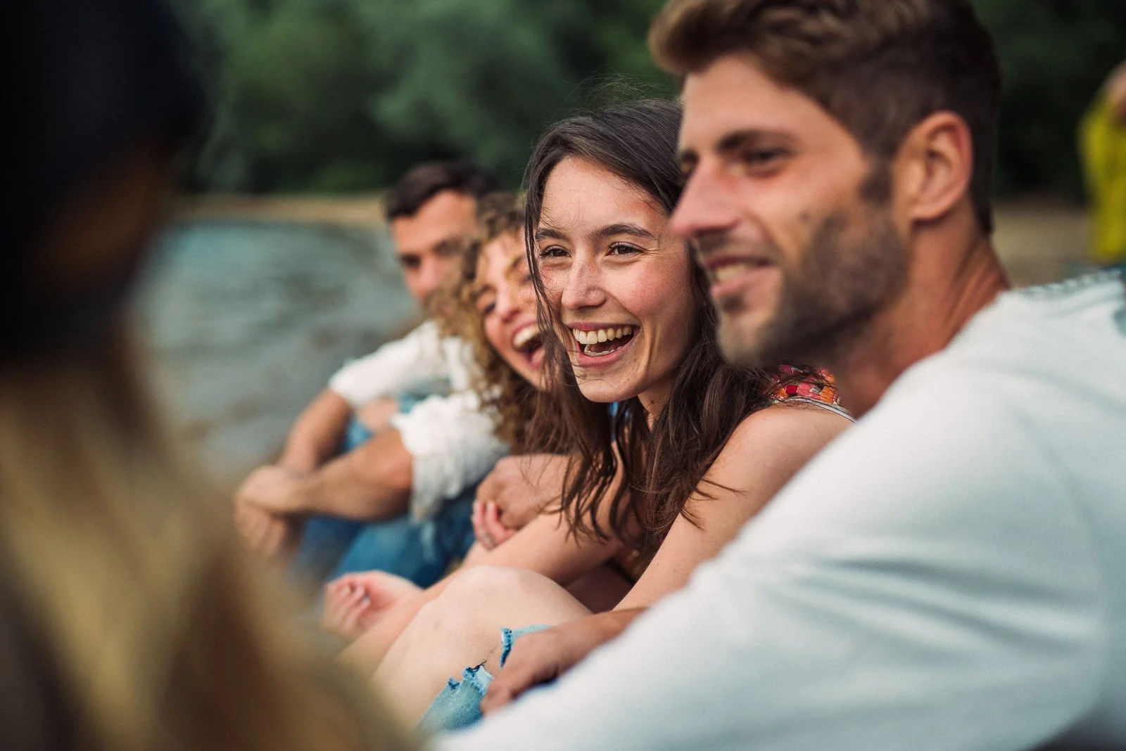 A group of friends sitting by a lake, smiling and laughing with each other.