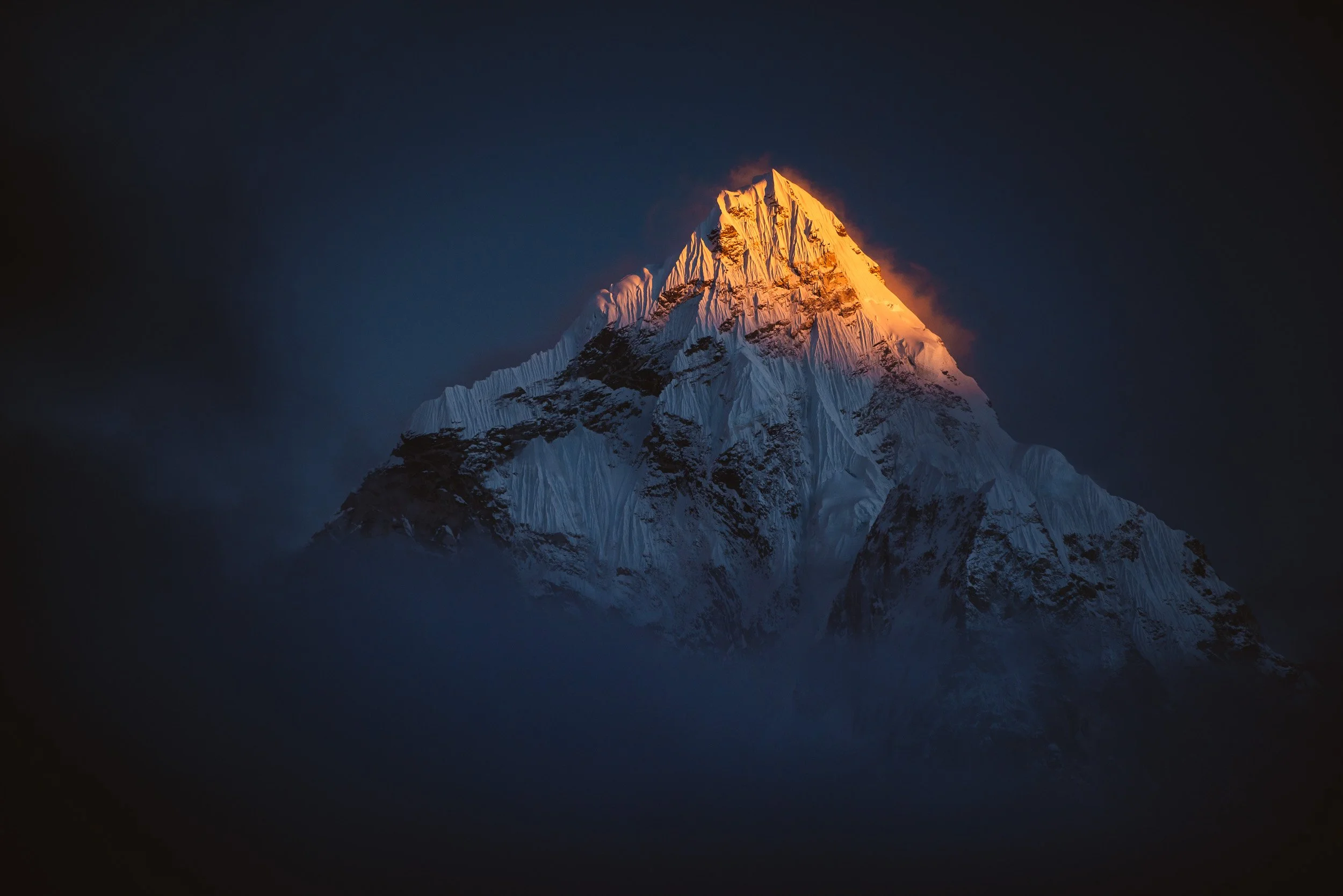 Snow-covered mountain peak illuminated by orange sunlight against a dark sky with clouds.