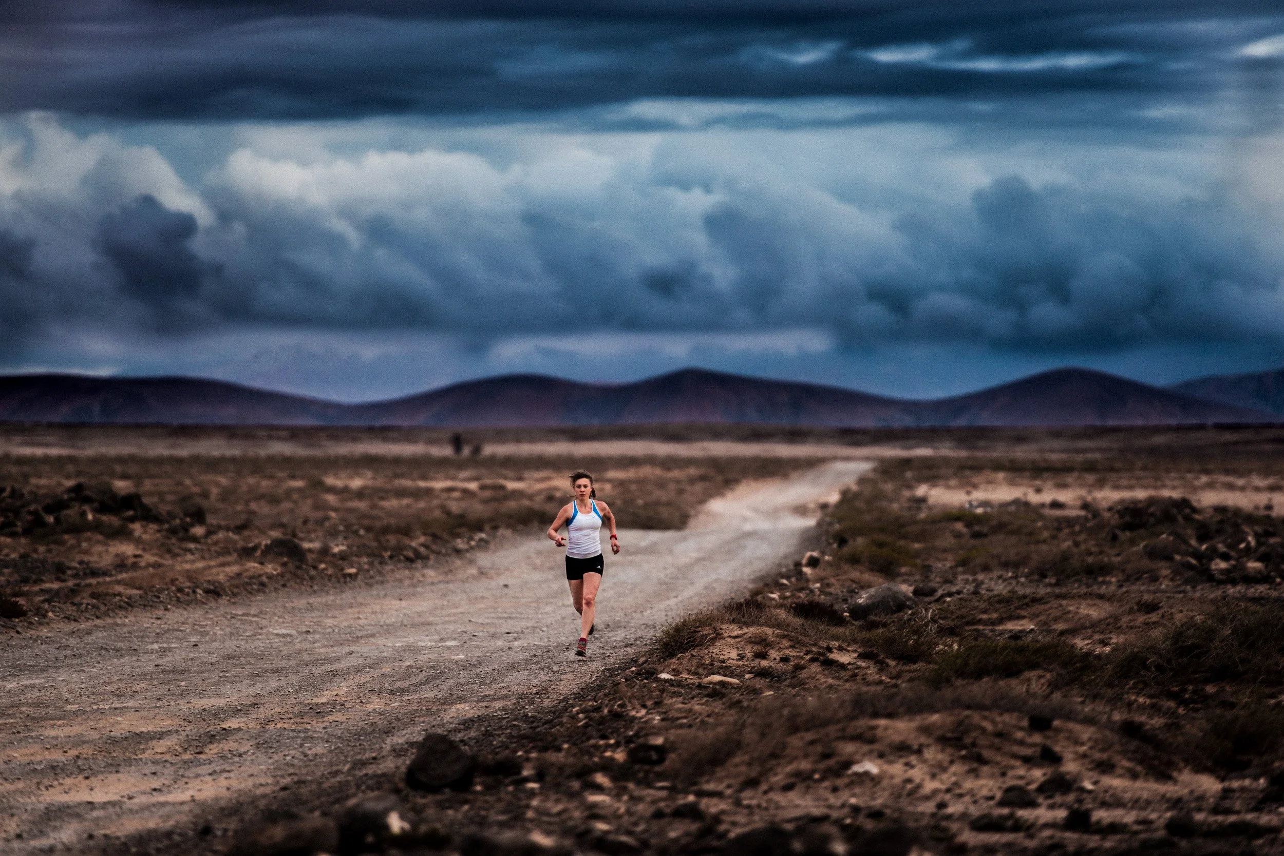 A woman jogging on a dirt trail in a desert landscape with mountains and dark clouds in the sky.
