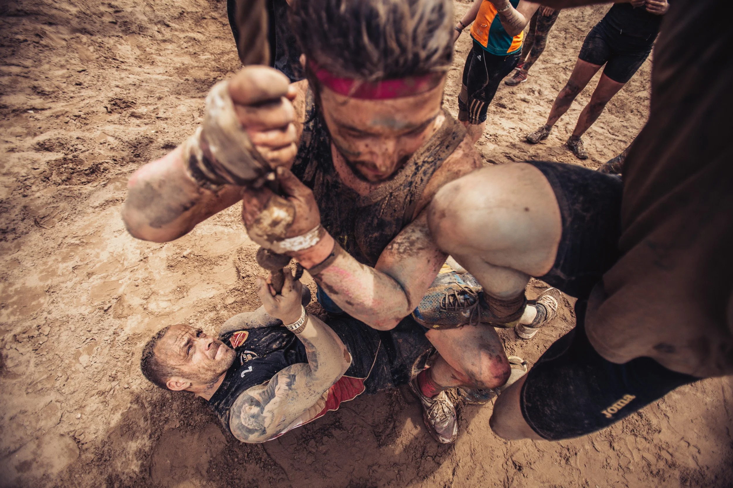 Two men involved in a muddy wrestling match on the ground, with muddy and dirt-covered attire and faces, while other people watch in the background.