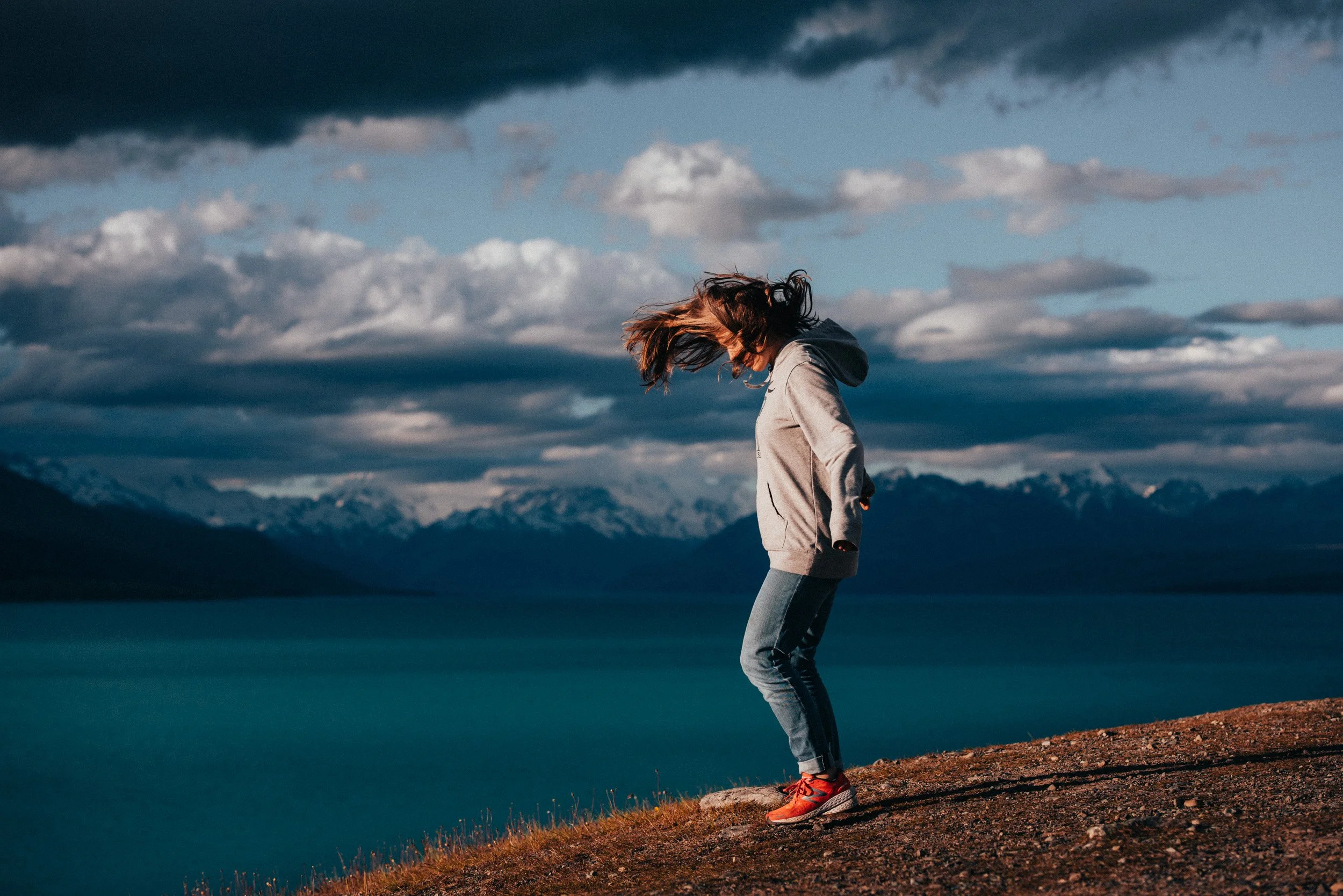 Woman in gray hoodie and jeans jumping near a body of water with mountains and cloudy sky in the background.