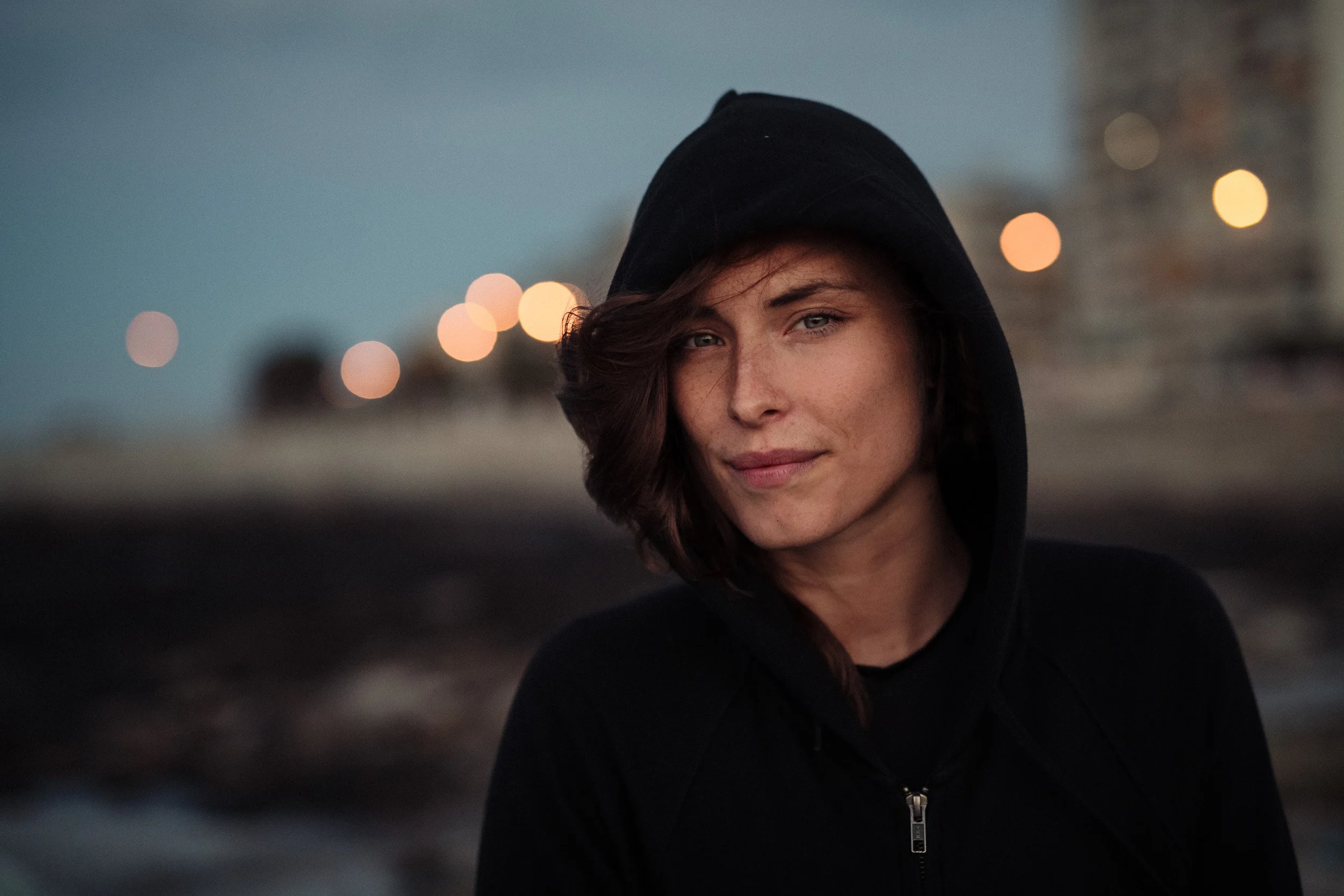 A young woman with short wavy brown hair, wearing a black hoodie, is outdoors during dusk with blurred city lights in the background.