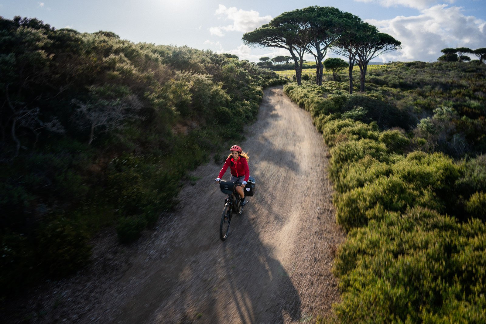 A person cycling on a dirt trail through a green, hilly landscape with trees and shrubs, under a partly cloudy sky.