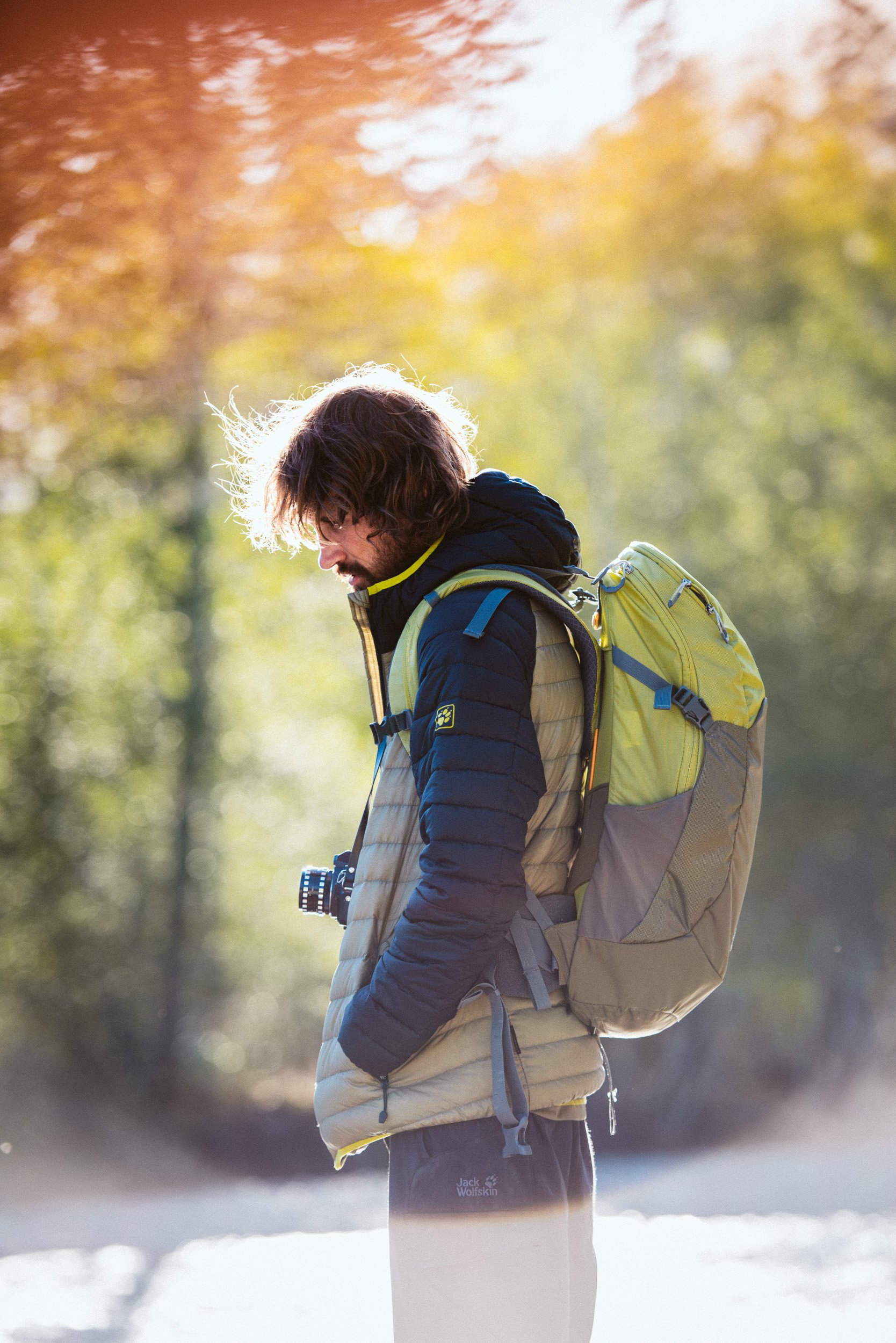 A man with shoulder-length hair and a beard stands outdoors in a forest, wearing a black and beige puffer jacket and a lime green and gray backpack, with a camera hanging around his neck, looking down.