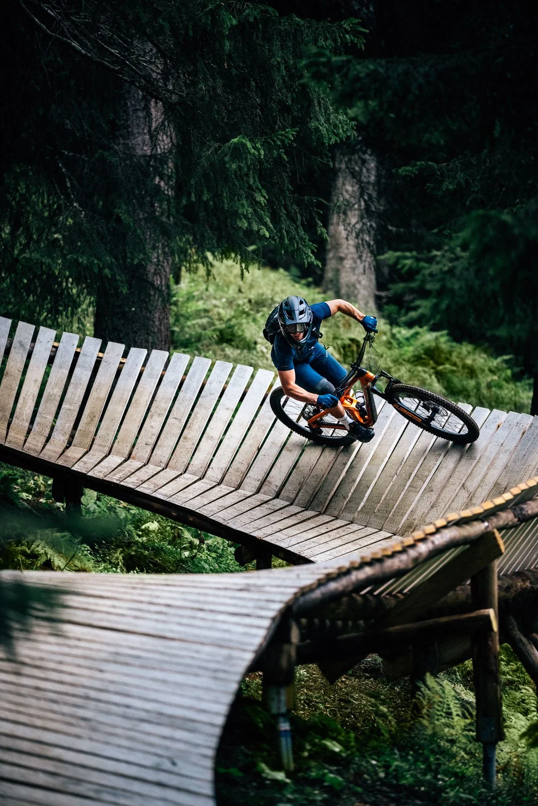 A mountain biker wearing a helmet and gloves navigates a steep wooden trail through a forest.