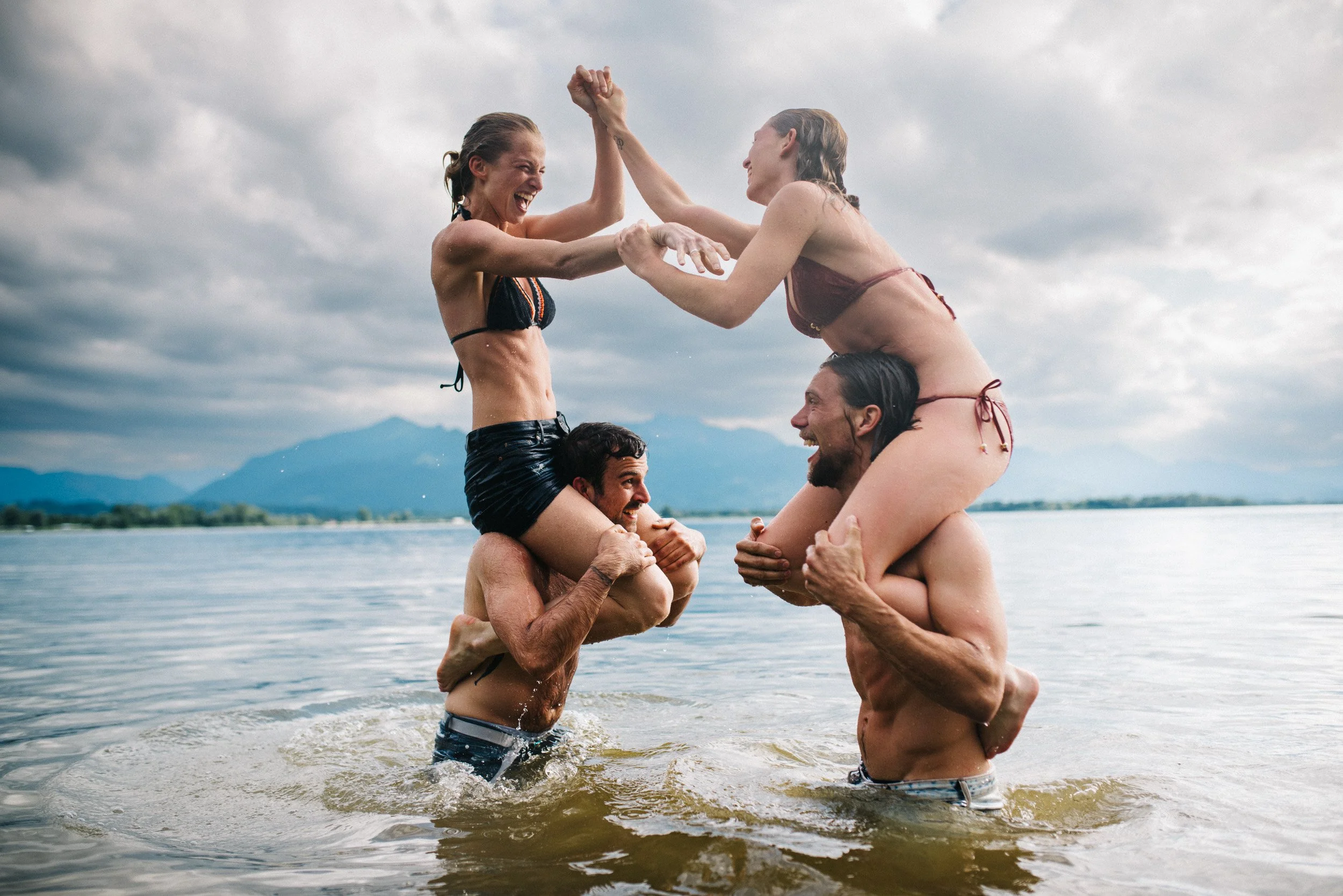 Four people are in the water, two men and two women, making a human pyramid. The women are on the men's shoulders, and they are holding hands and smiling. The background shows a lake, mountains, and a cloudy sky.