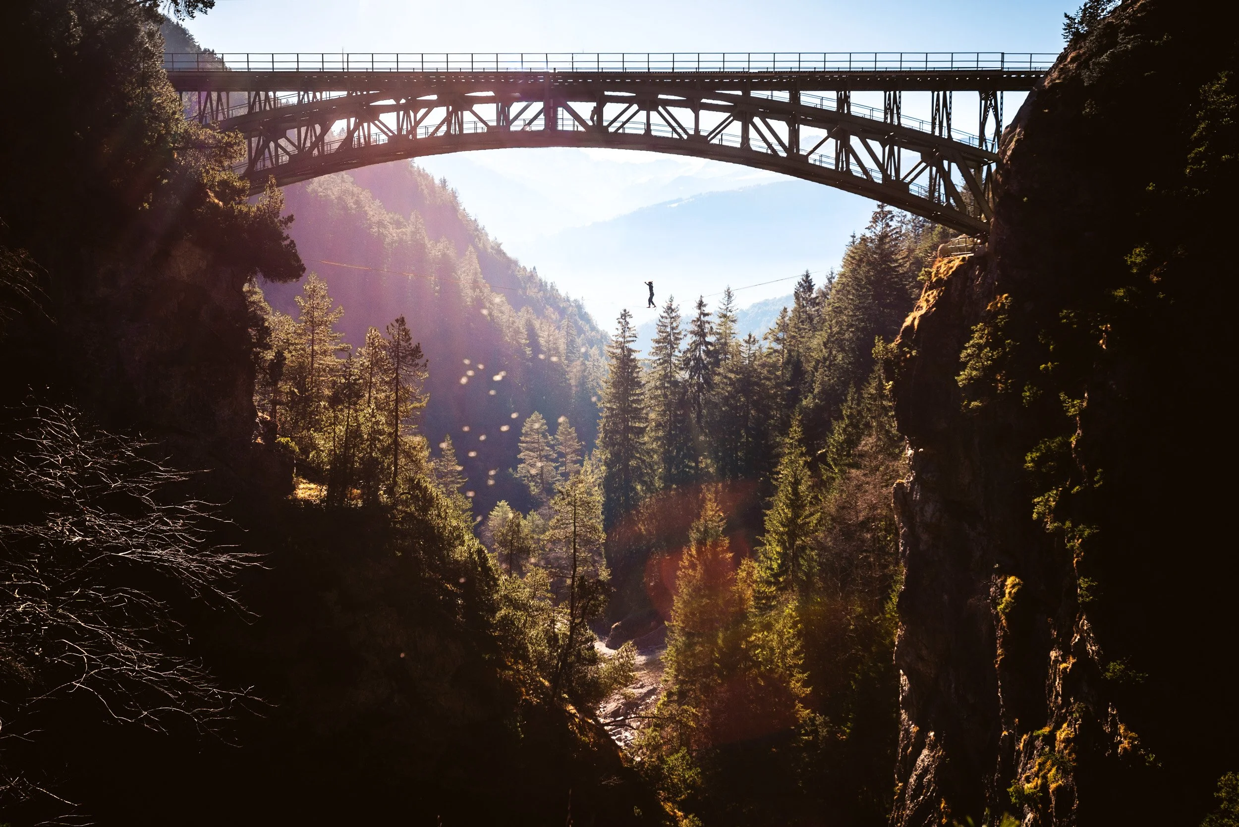 A person walking on a tightrope between two cliffs, with a bridge overhead, in a mountainous forested area.