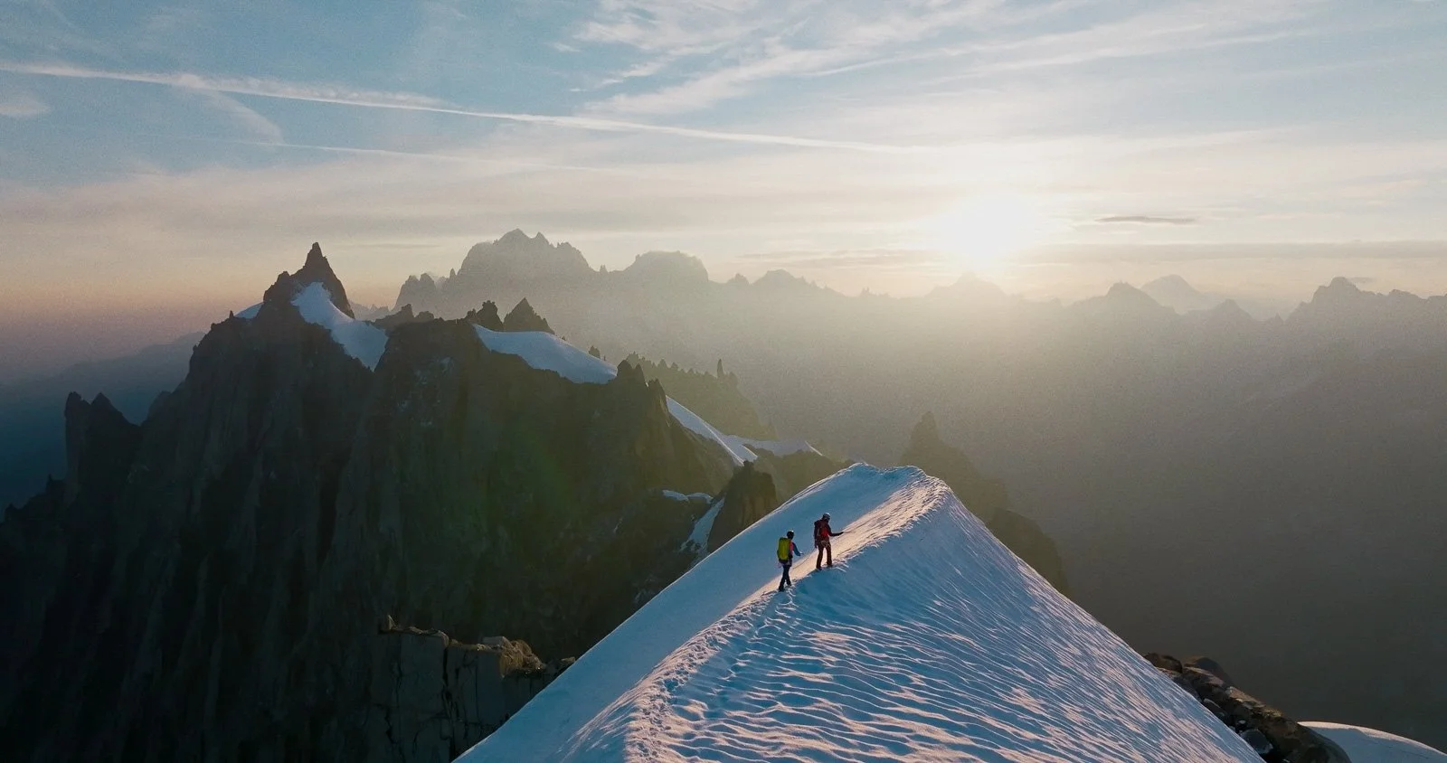 Two climbers traverse a snowy ridge on a mountain at sunrise, with jagged peaks and a cloudy sky in the background