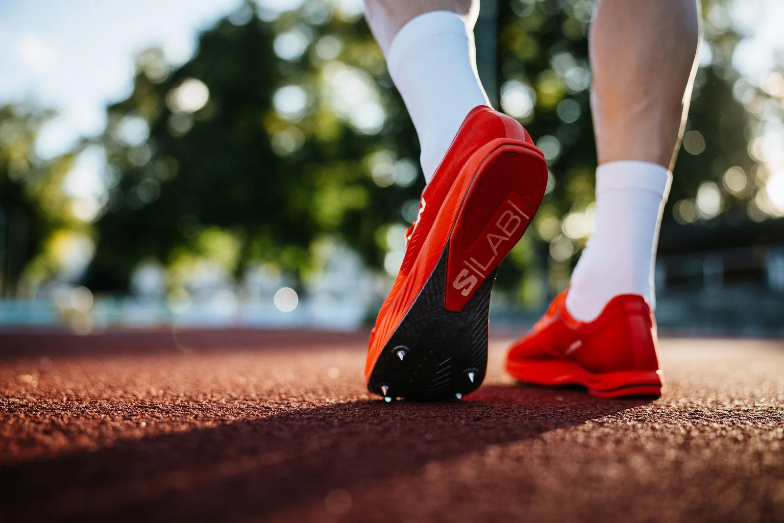 A person wearing white socks and red cleats standing on a running track with one foot slightly lifted, with a focus on their athletic shoes and legs, blurred green trees and sunlight in the background.