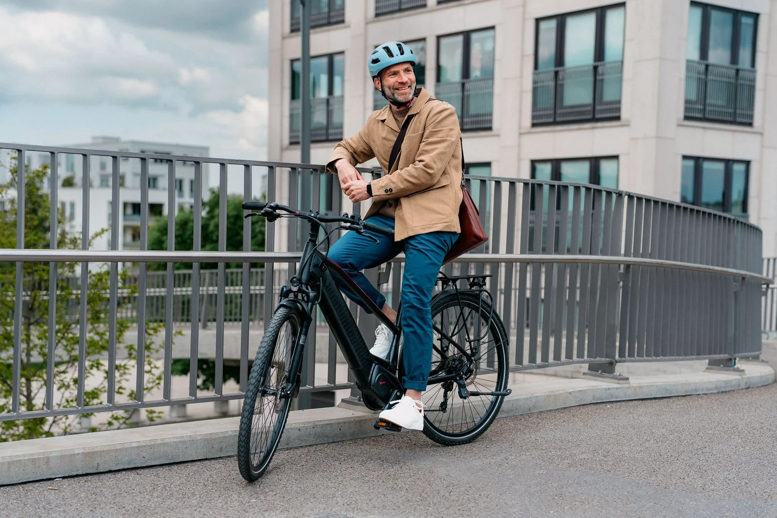 A man wearing a blue helmet, a tan jacket, and blue pants is sitting on an electric bike in an urban area with modern buildings in the background. He is smiling and looking to the side.