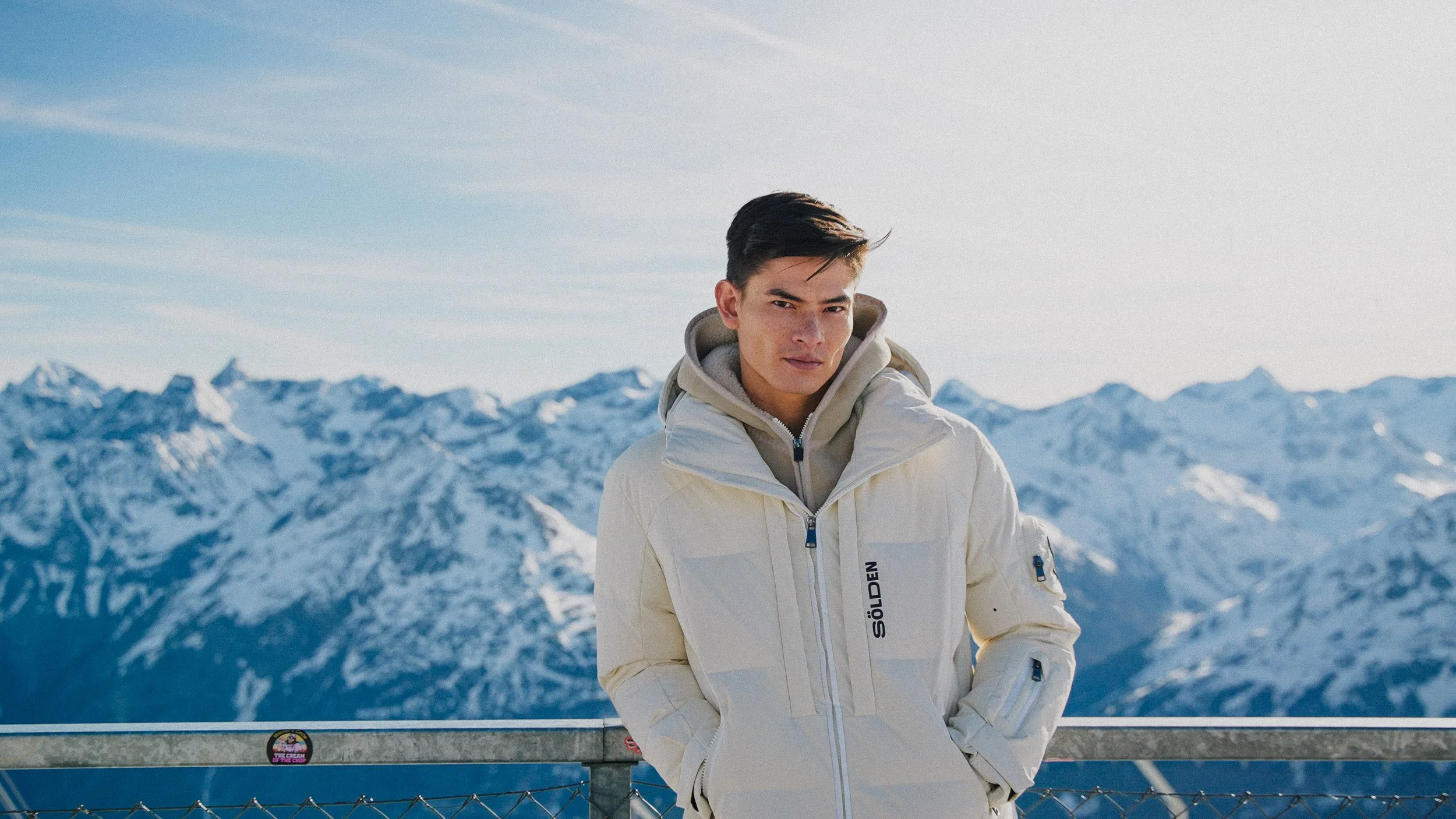 A young man standing outdoors with snow-covered mountains in the background, wearing a white winter jacket with his hands in his pockets.