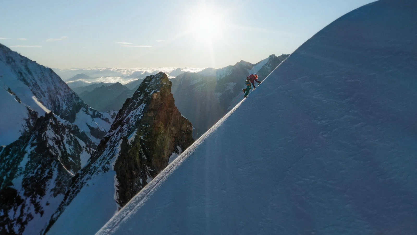 A person in a red jacket and black pants climbing a snowy mountain slope with a rope, surrounded by snow-covered peaks under a sunny sky.