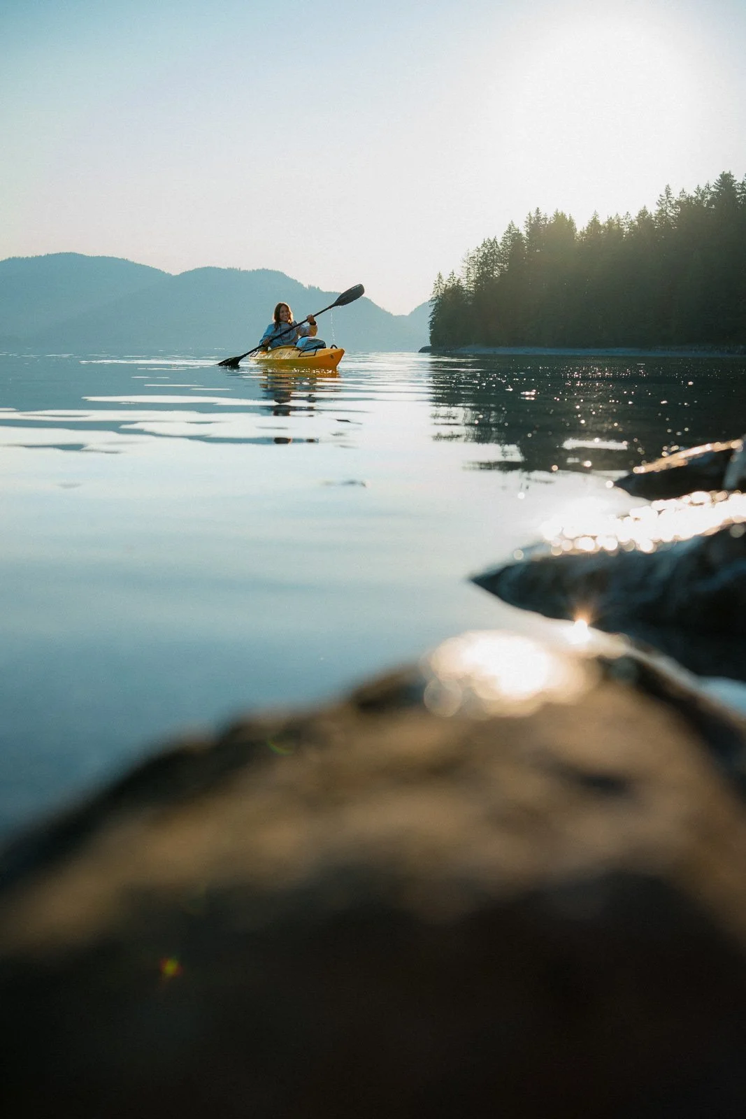 A person kayaking on a calm lake with mountains and trees in the background, near a rocky shoreline.