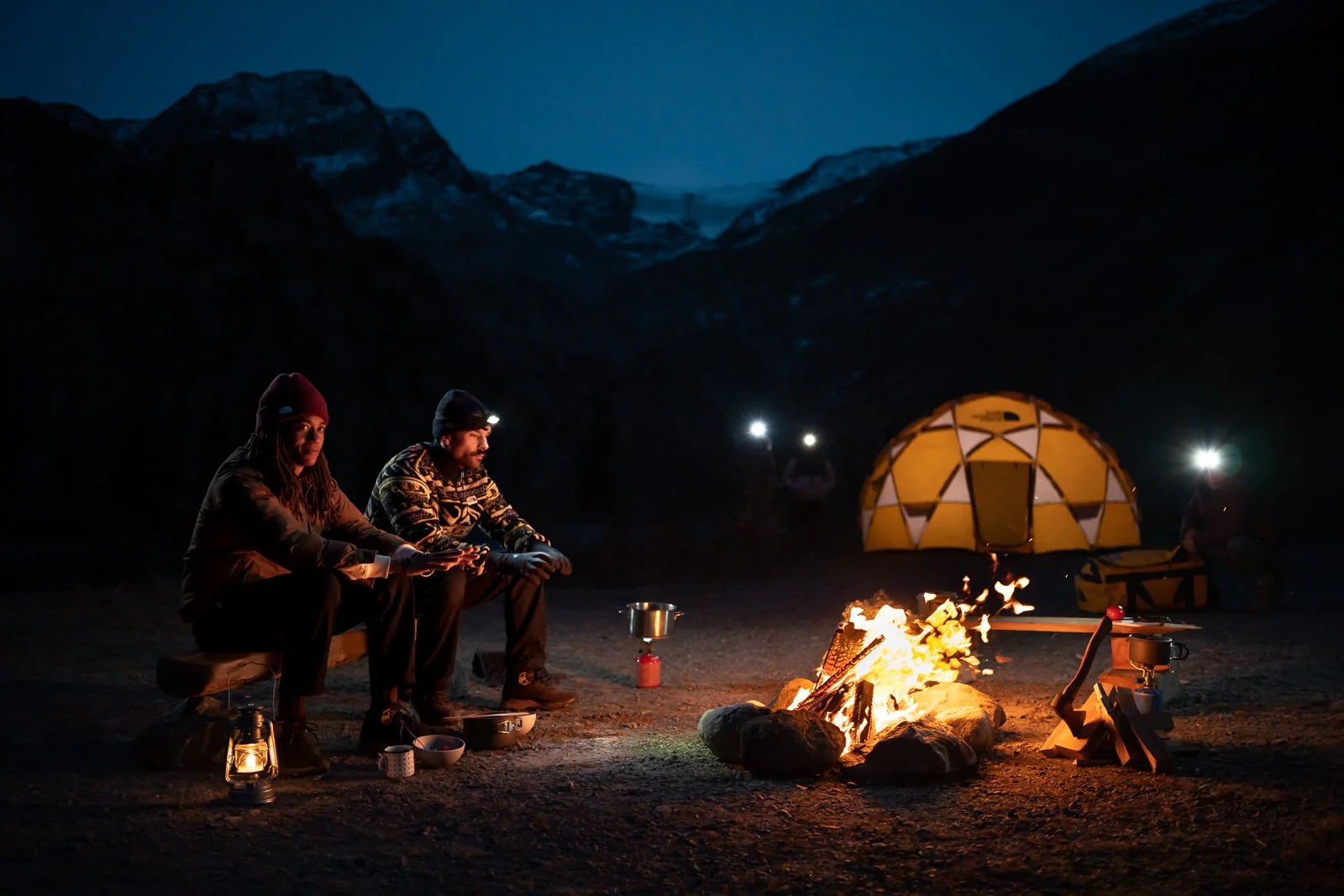 Two people sitting around a campfire at night in a mountainous landscape, with a tent and other camping gear visible.