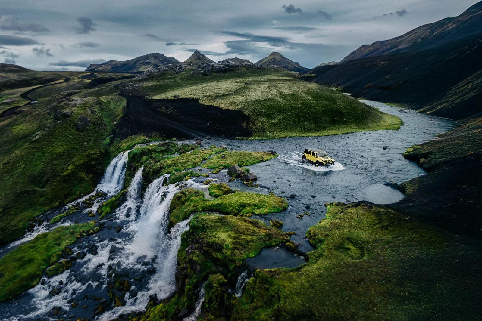 A yellow off-road vehicle driving through a river in a lush, green valley with waterfalls and mountains under a cloudy sky.