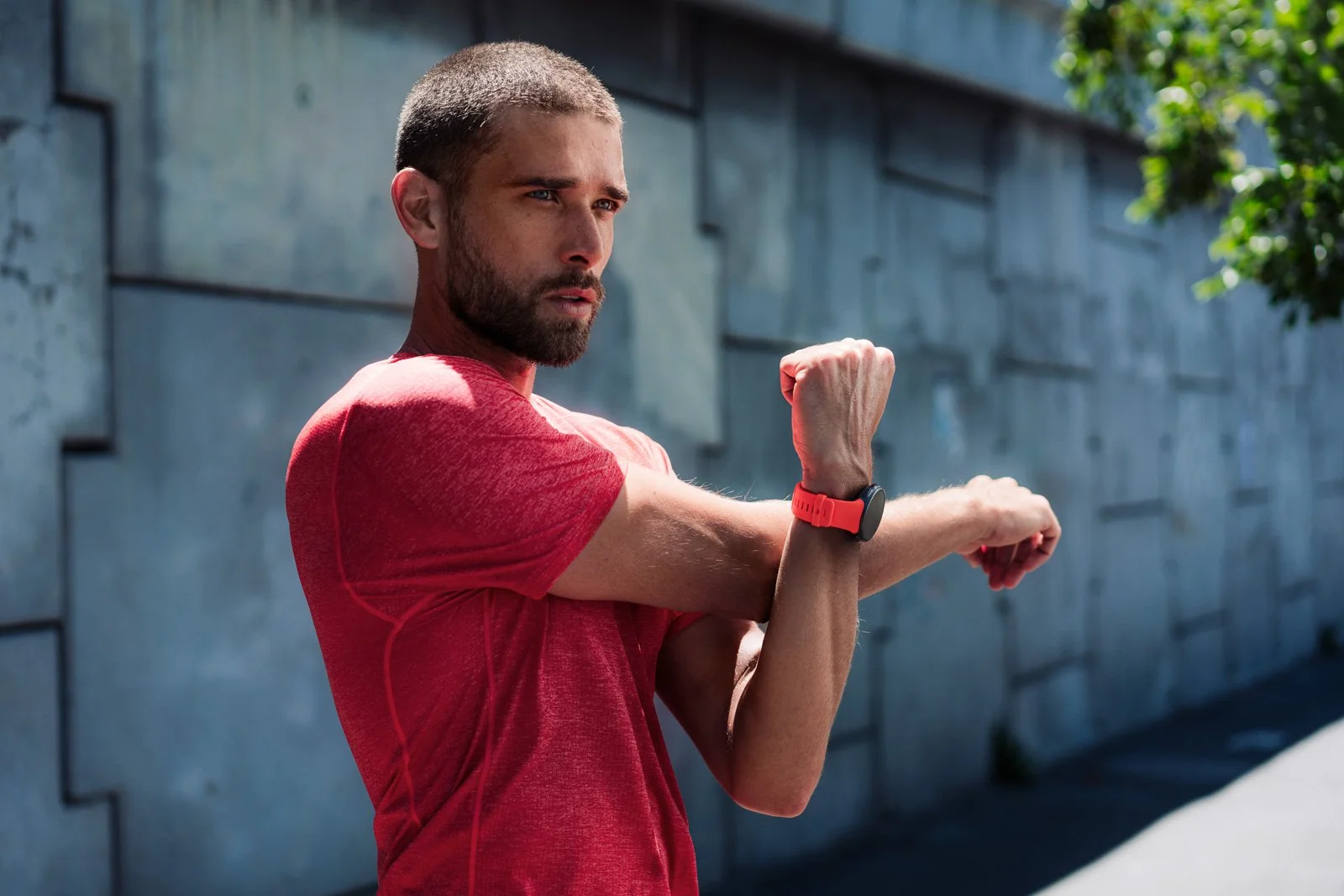 A man in a red athletic shirt checks his fitness tracker while stretching outdoors next to a concrete wall.