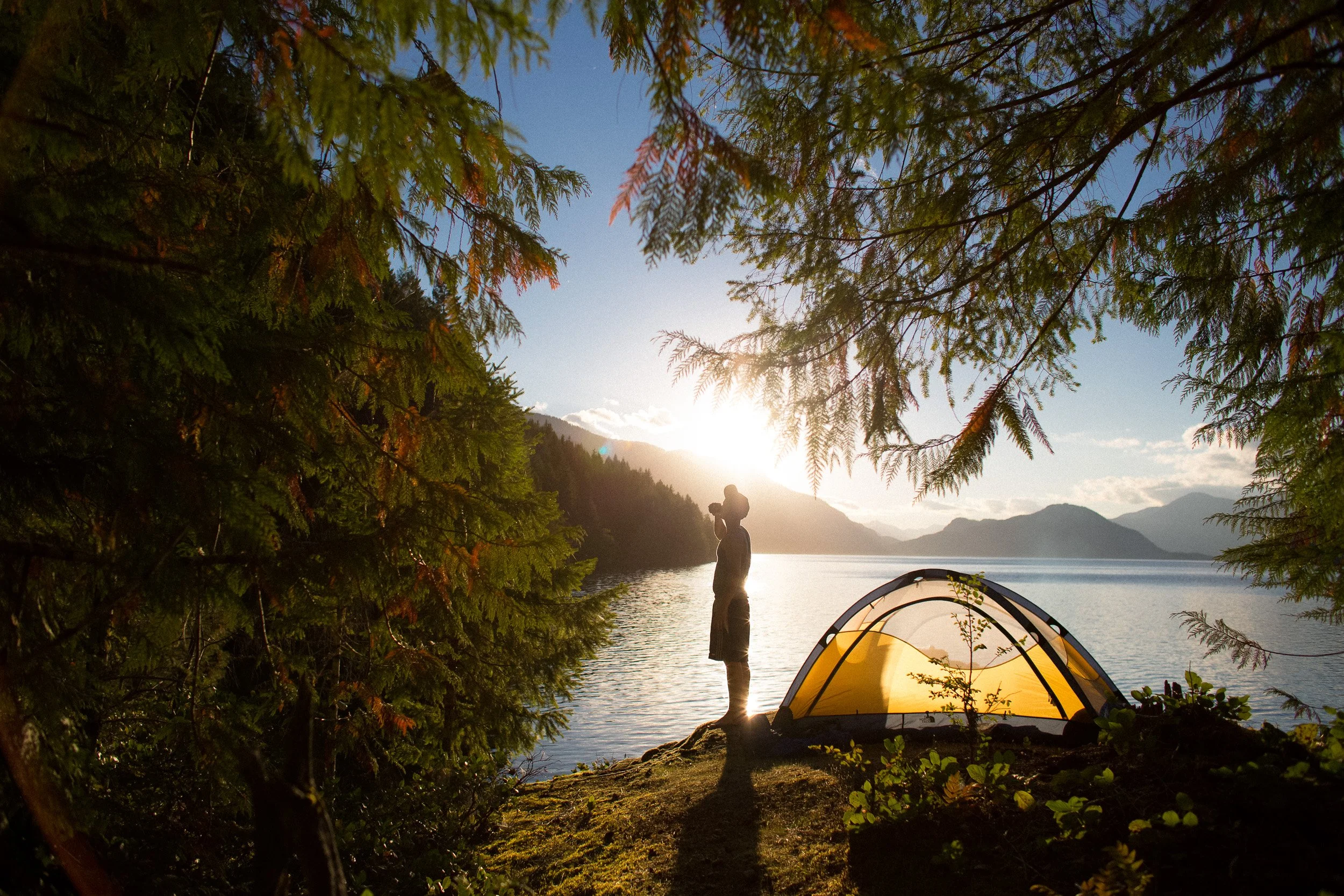 Person standing near tent by a lake at sunset, surrounded by trees and mountains in the background.