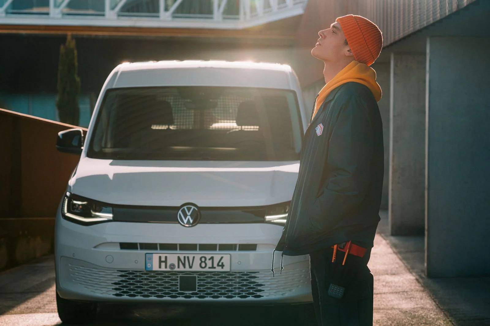 Young man with short hair and a fair complexion standing in front of a white Volkswagen van, wearing an orange beanie, a yellow hoodie, and a dark jacket with a small logo. The man is facing sideways with his eyes closed, basking in sunlight.
