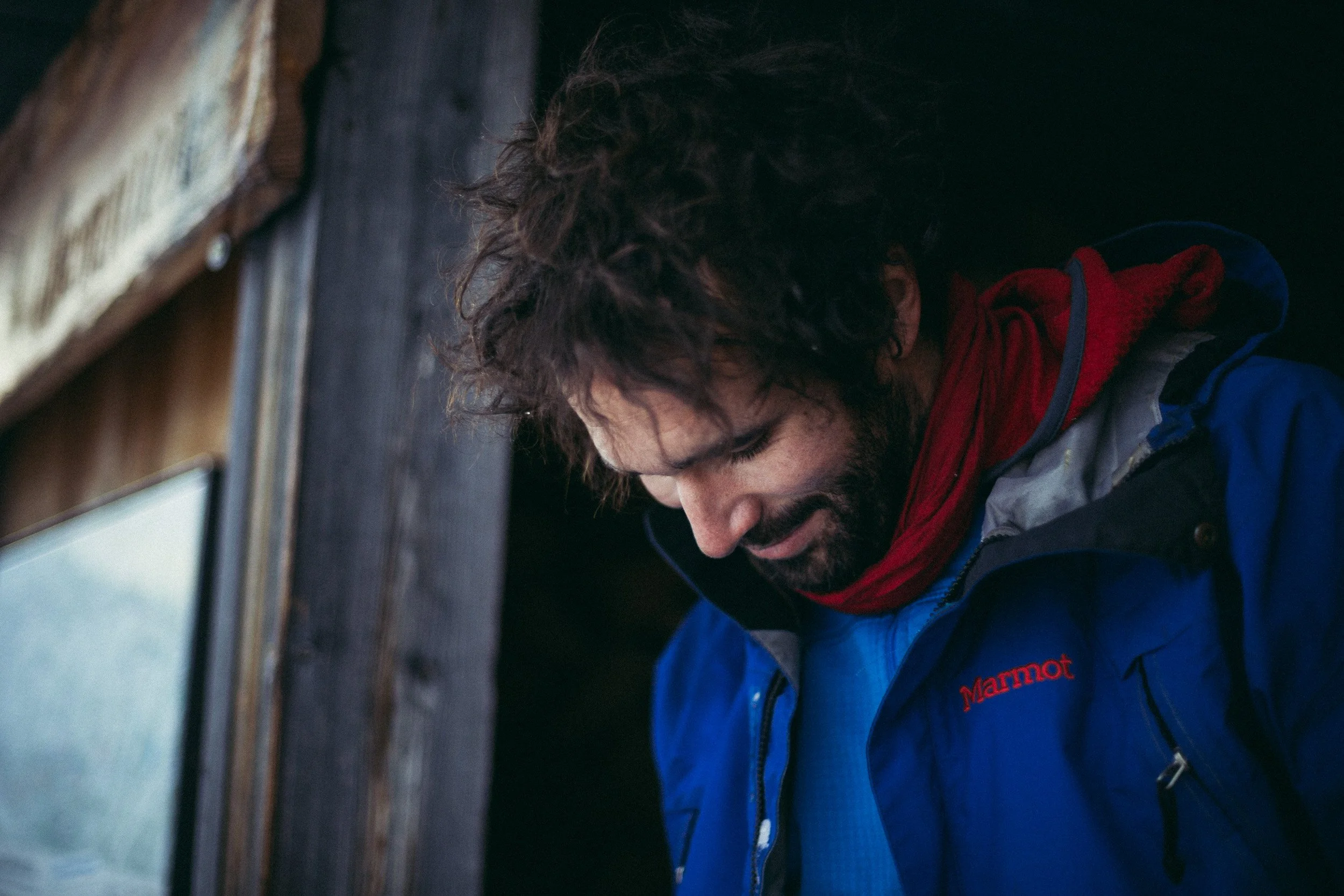 A man with curly hair and a beard looking downward, wearing a blue Marmot jacket and a red scarf, standing in front of a wooden structure.