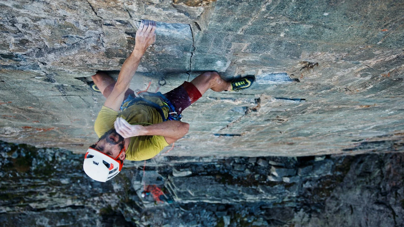 A man rock climbing on a steep outdoor rock face, wearing a helmet, climbing harness, and athletic clothing.
