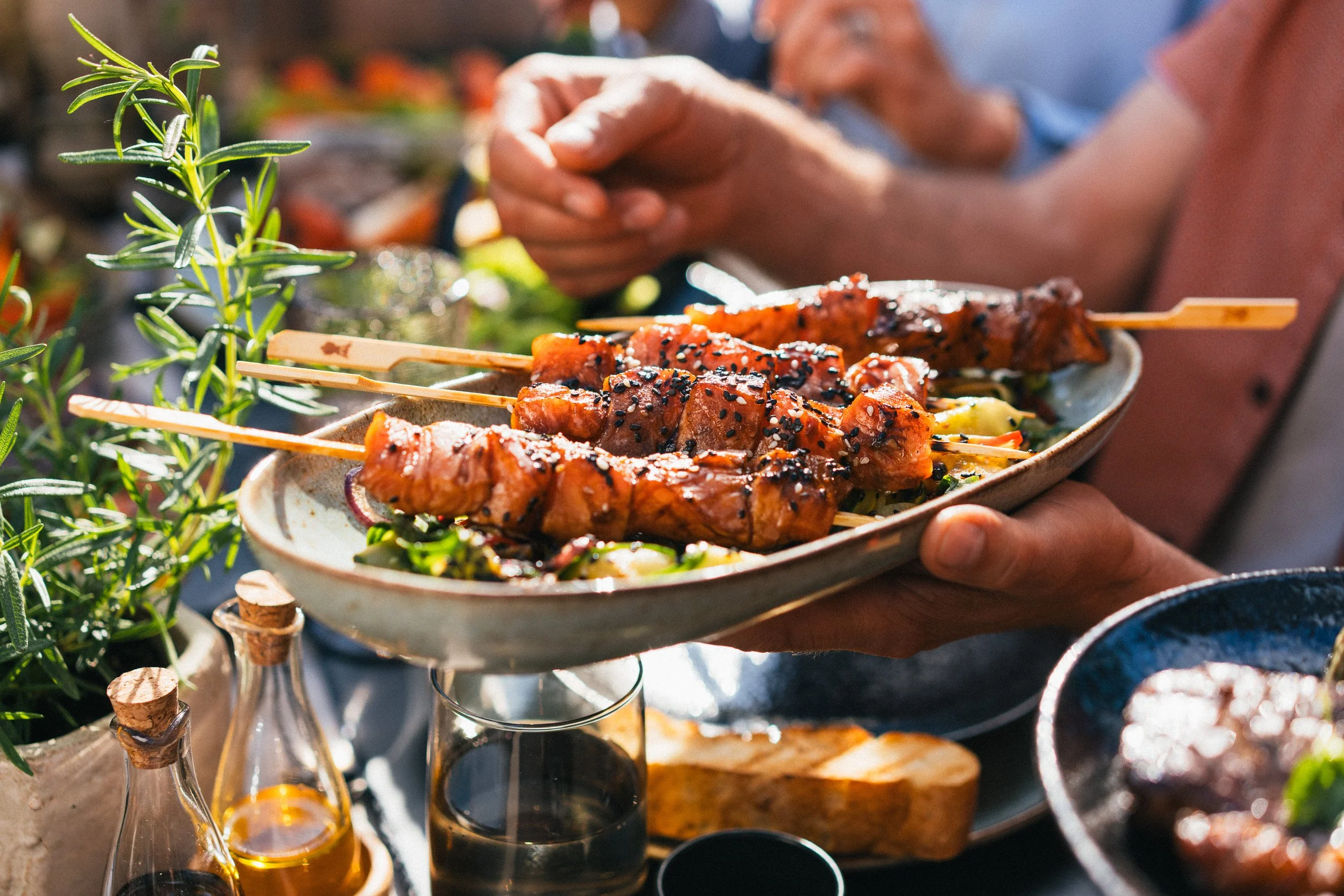 Plate of skewered grilled meat garnished with sesame seeds, held by a person with a background of other dishes and greenery.