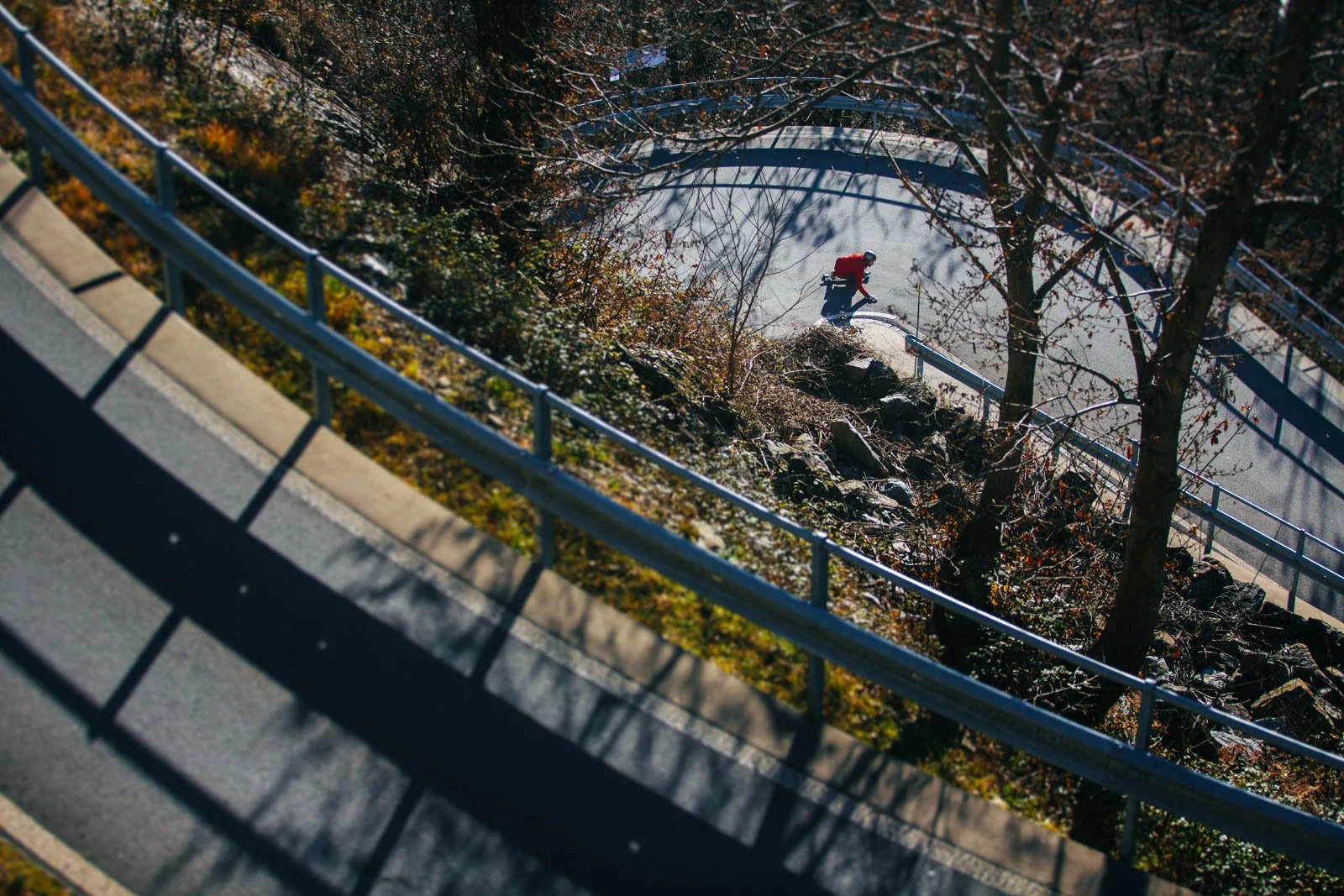A person in a red jacket and helmet walking along a concrete pathway, bordered by metal railings, with trees and rocks below, seen from above on a sunny day.