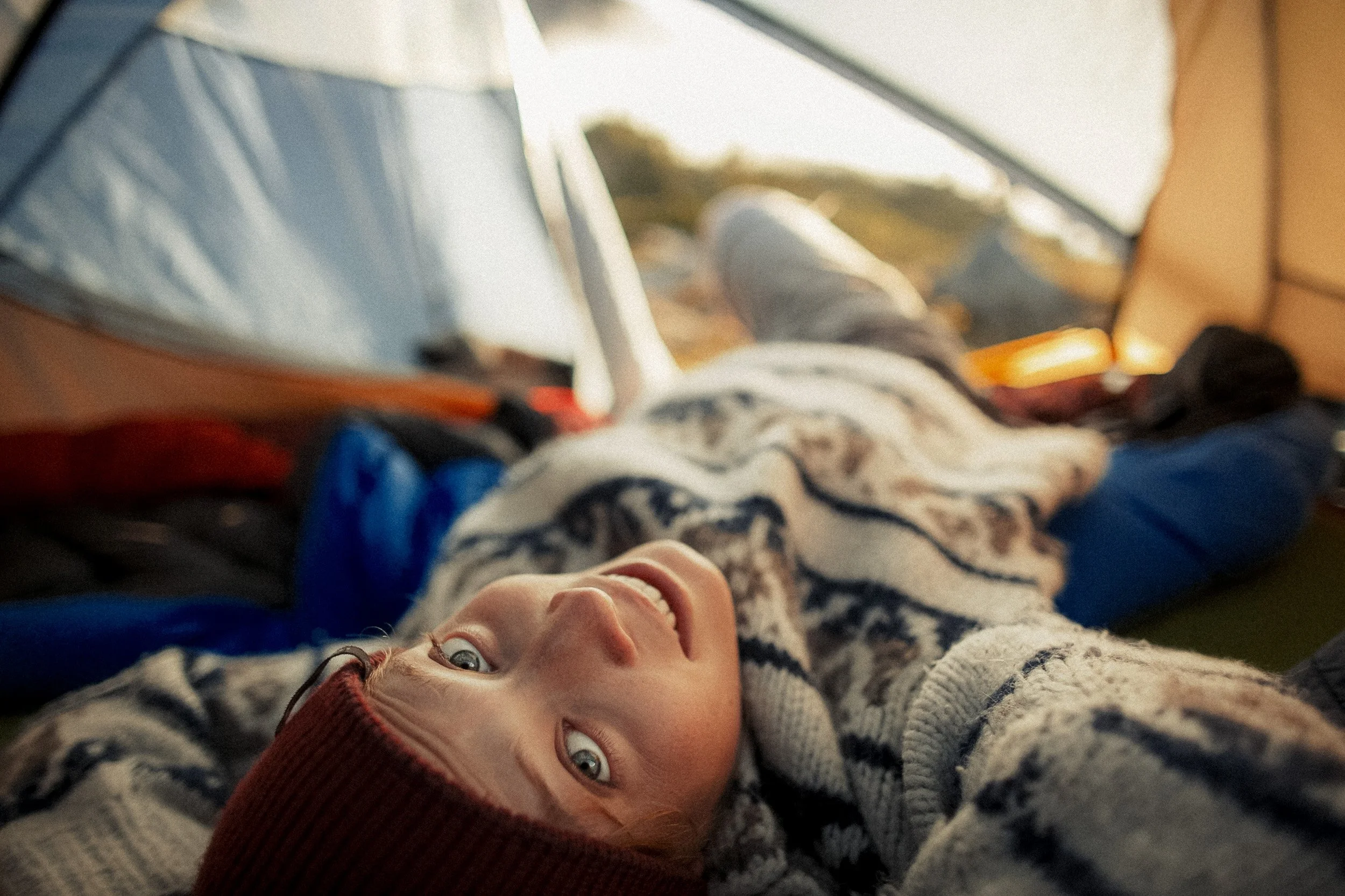 Person lying inside a tent with a patterned sweater and beanie, looking up at the camera with a smile. Camping supplies and outdoor scenery are visible.