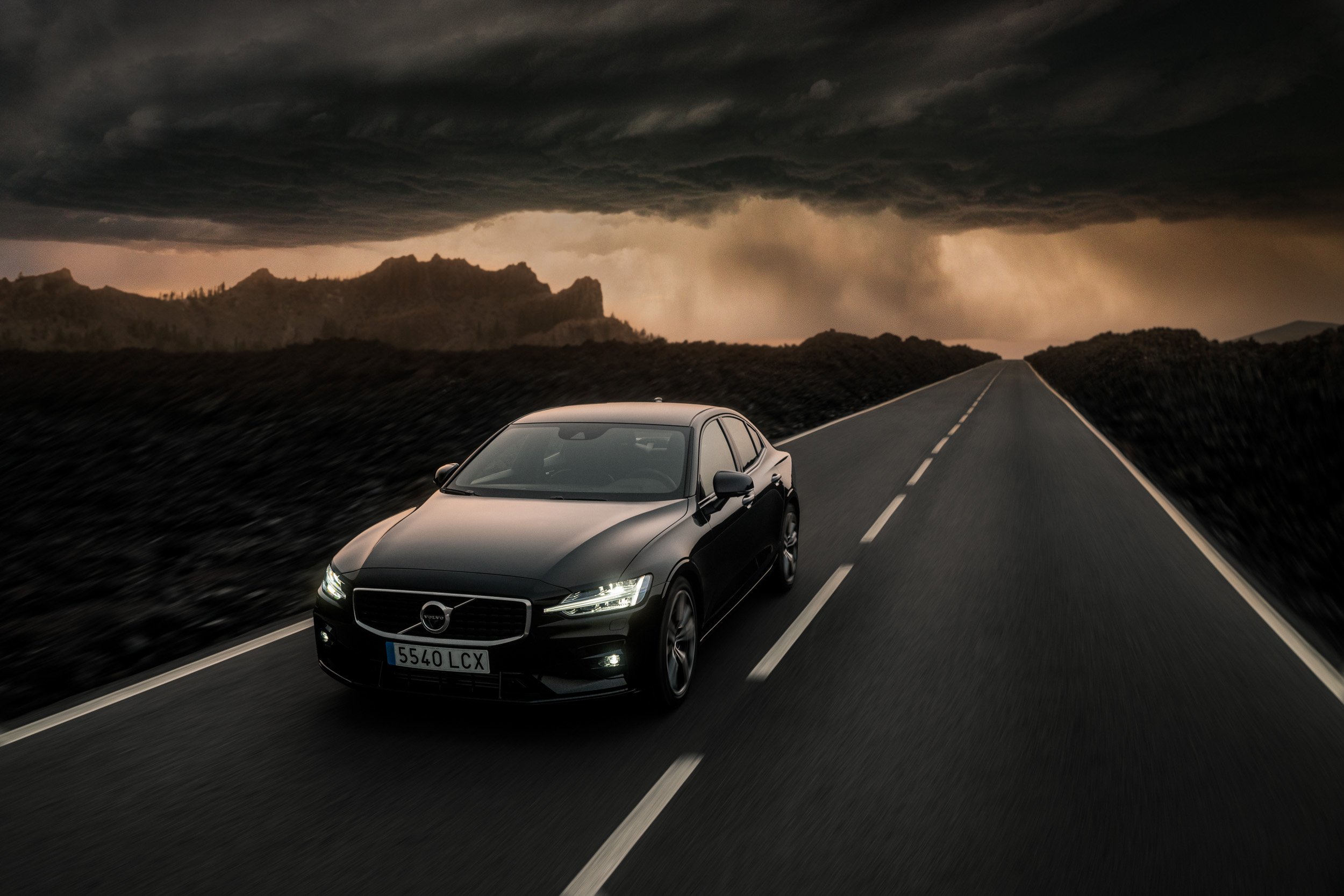 A black Volvo sedan driving on a two-lane road under dark, stormy clouds.