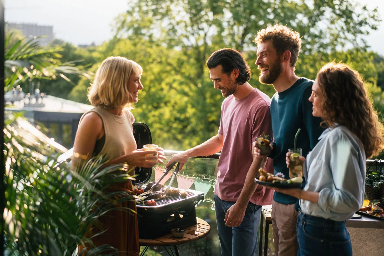 A group of four friends enjoying a backyard summer barbecue outdoors, with two men and two women smiling and holding drinks, as the woman grills food on an outdoor grill.