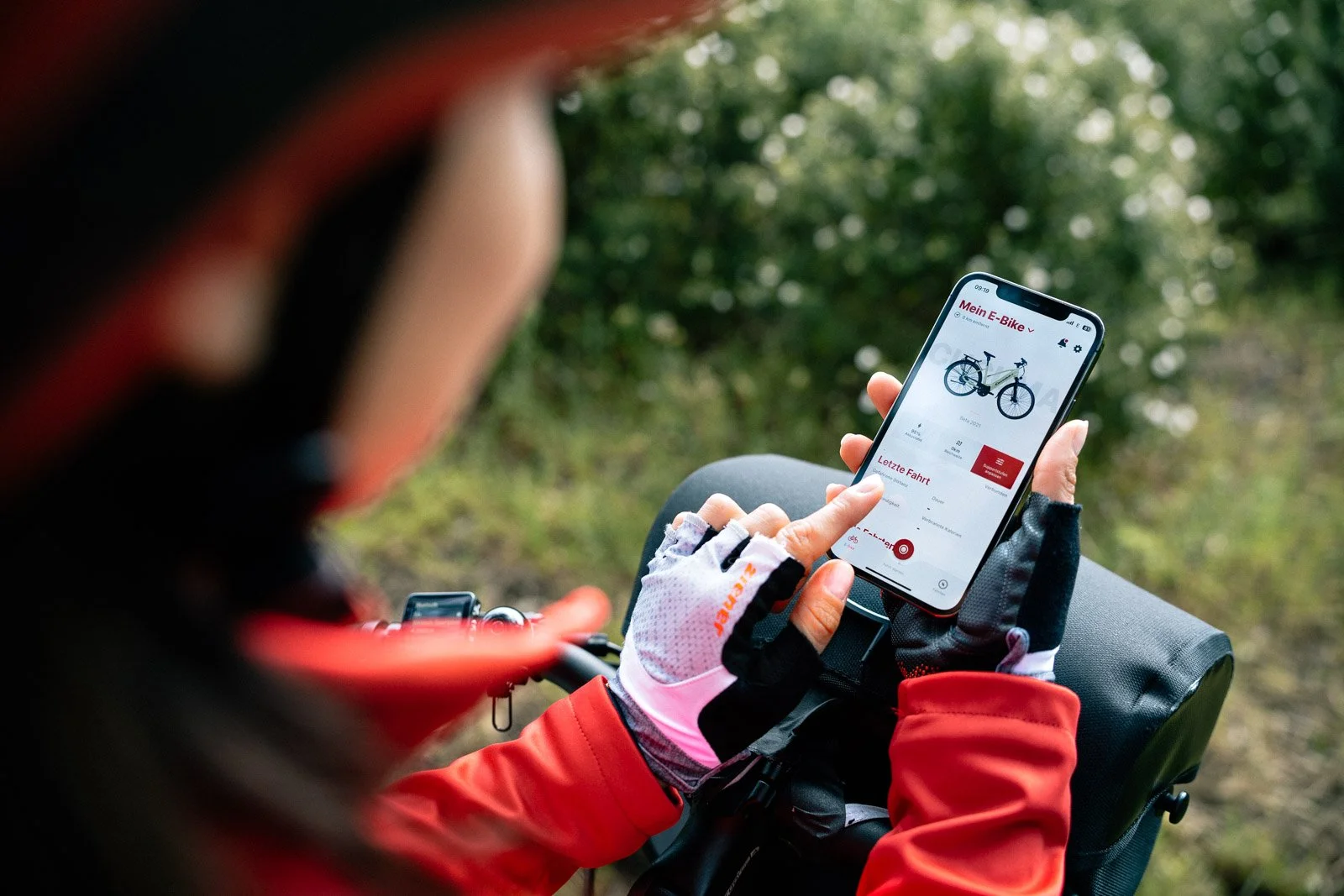 Person in cycling gear, wearing gloves, using a smartphone to select a bicycle on a bike rental app outdoors in a wooded area.