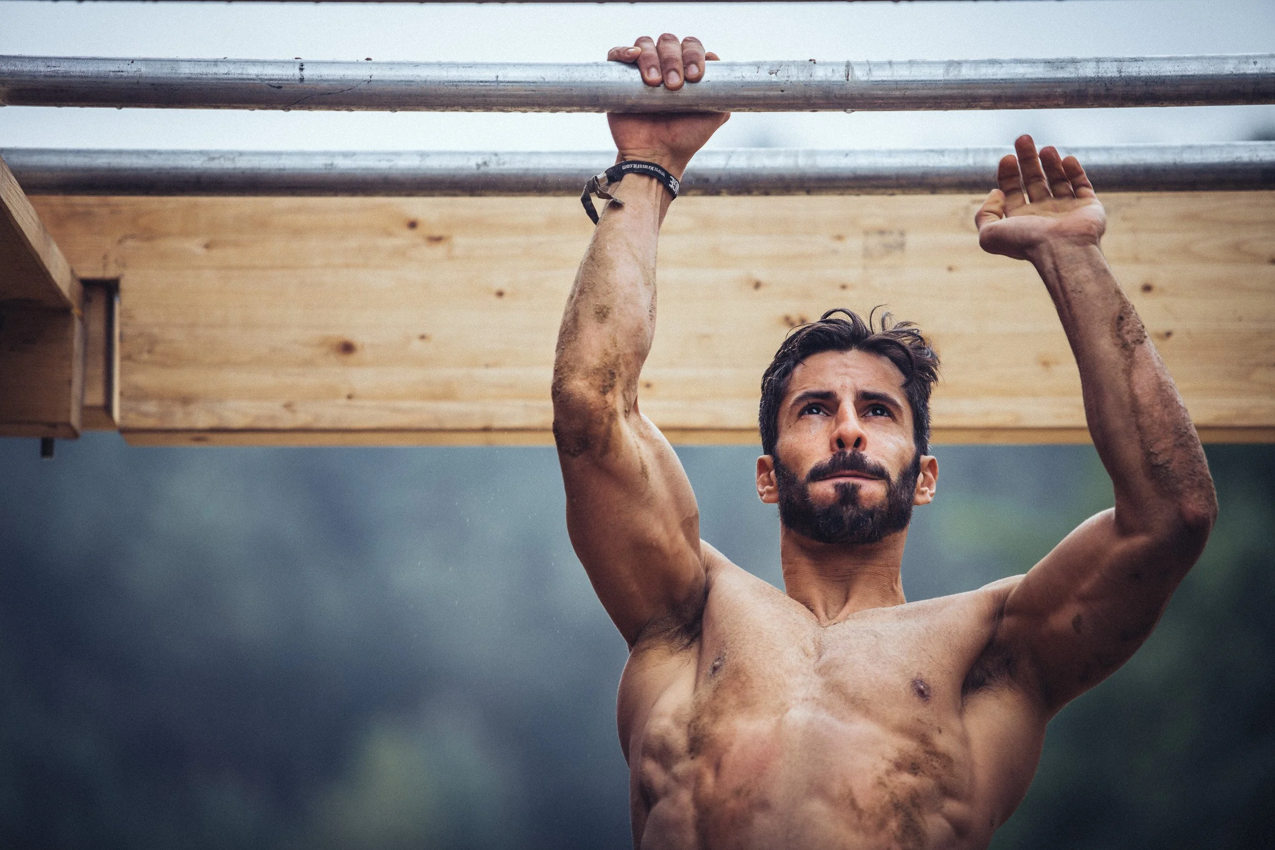 A shirtless man with a beard and dirty arms hanging from a wooden structure during an obstacle course race.