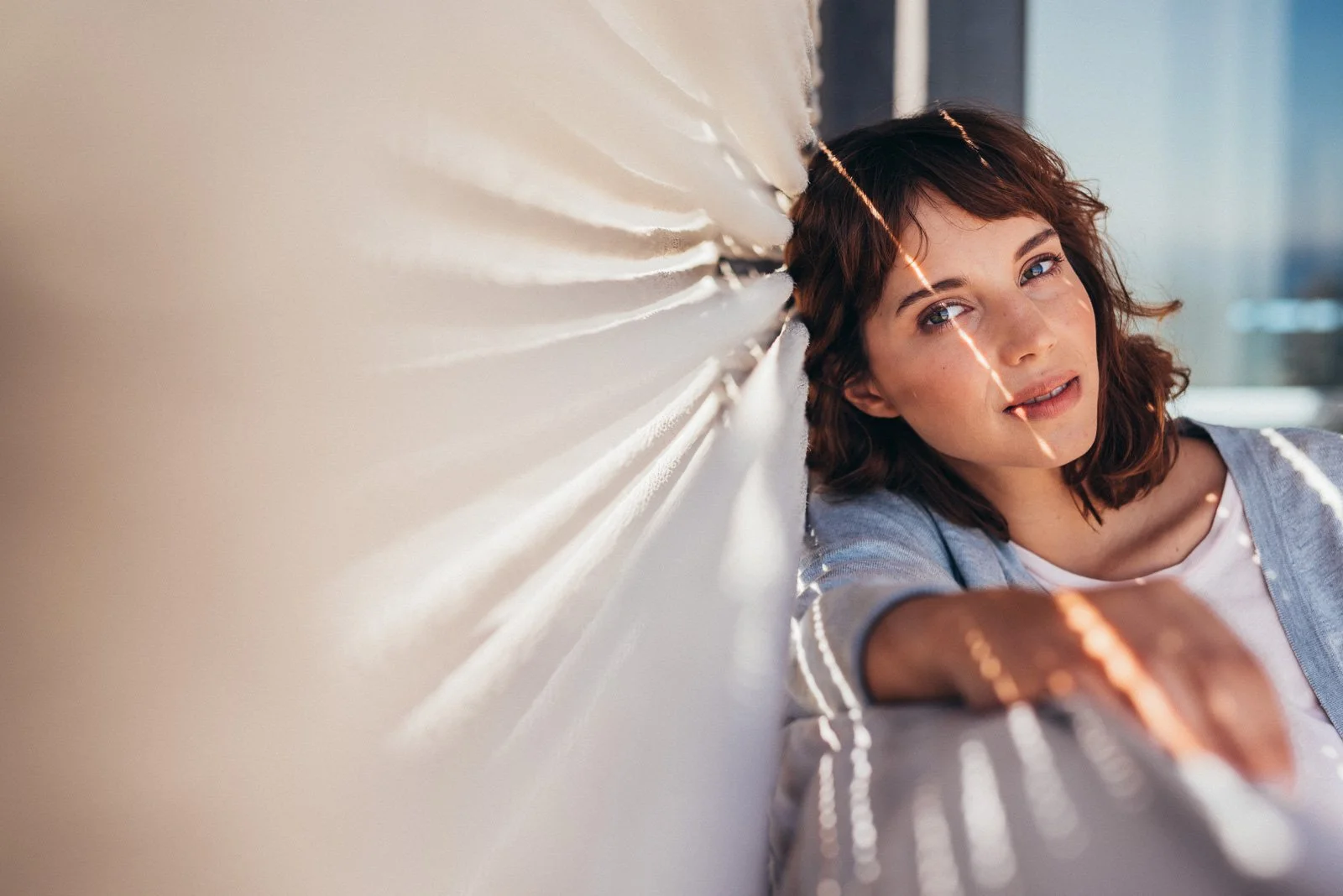 A young woman with short, wavy brown hair and light skin leaning against a wall, with sunlight creating diagonal lines across her face and arm.