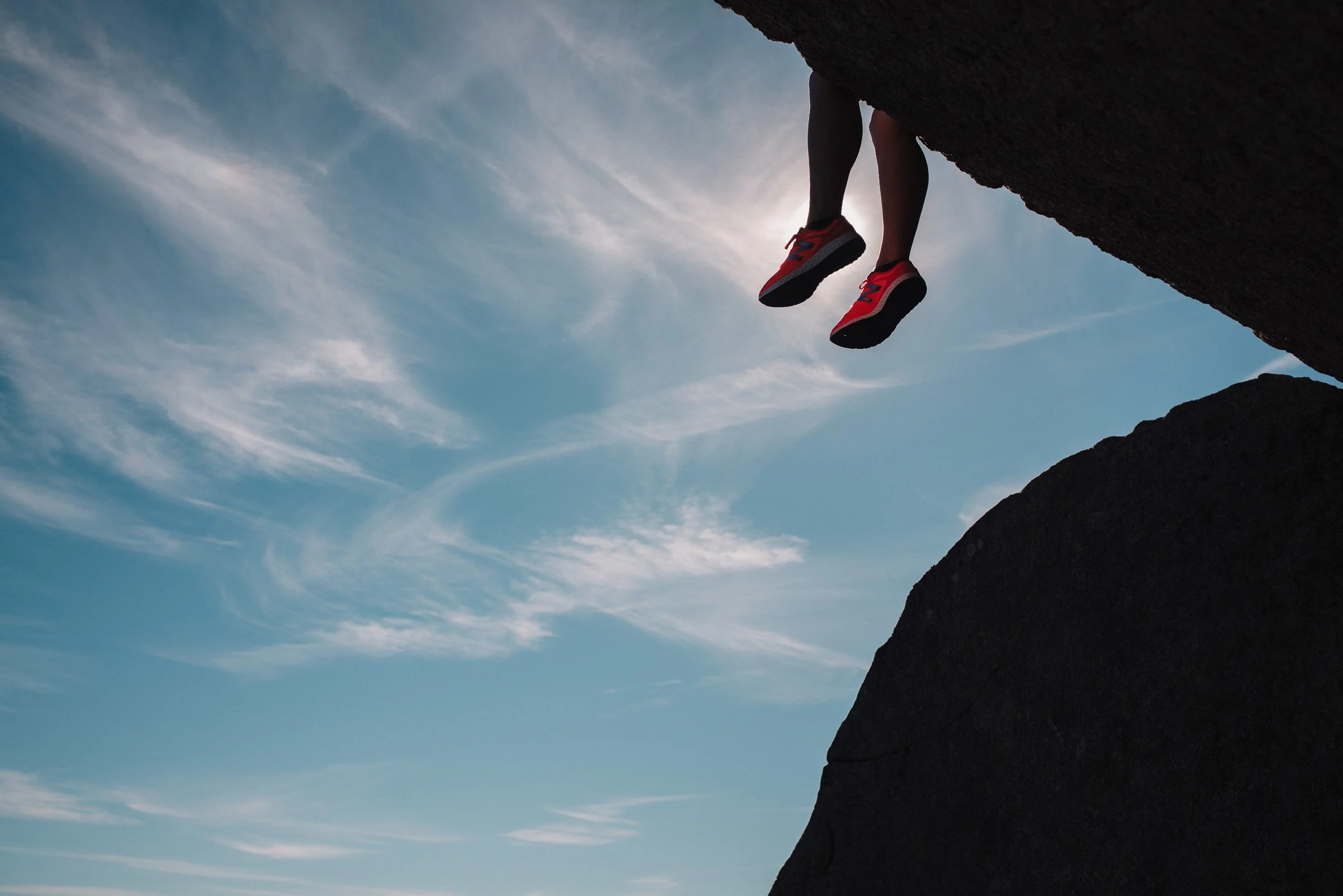 A person climbing a rock formation with only their legs and feet visible, wearing red and black sneakers, against a blue sky with wispy clouds.
