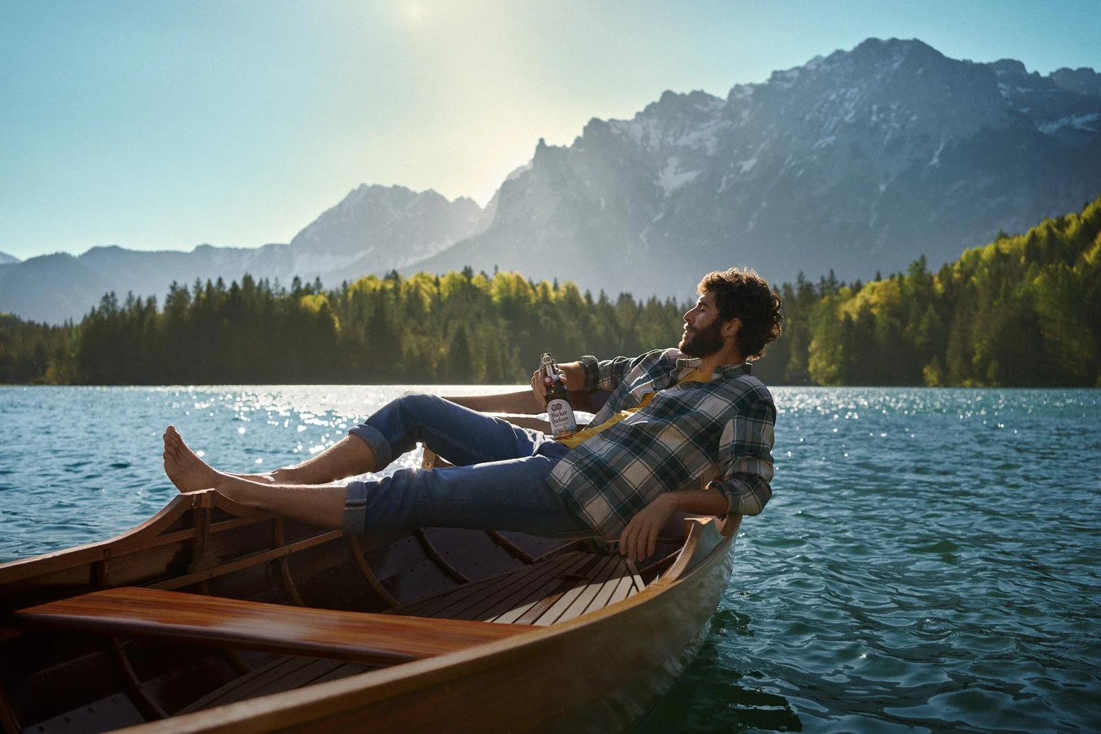 A man relaxing in a wooden rowboat on a lake with mountains and forest in the background, holding a beer.