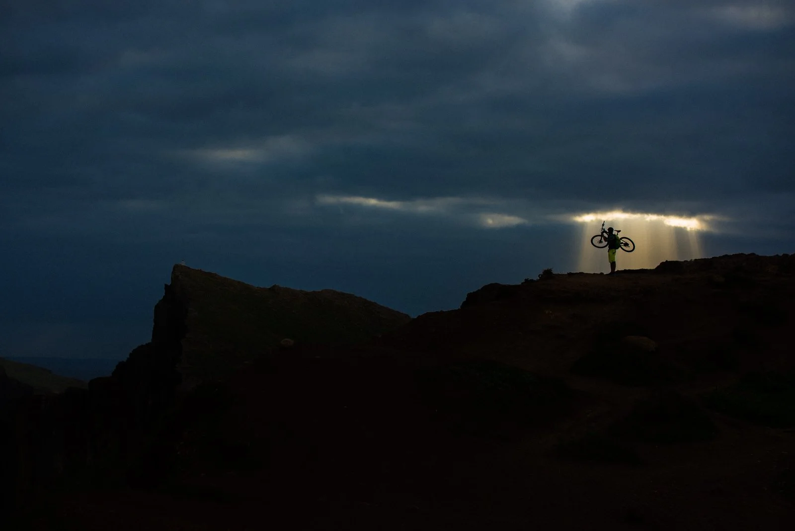 A person holding a bicycle over their shoulder standing on a mountain ridge during dusk or dawn, with dark clouds overhead and sunlight breaking through.