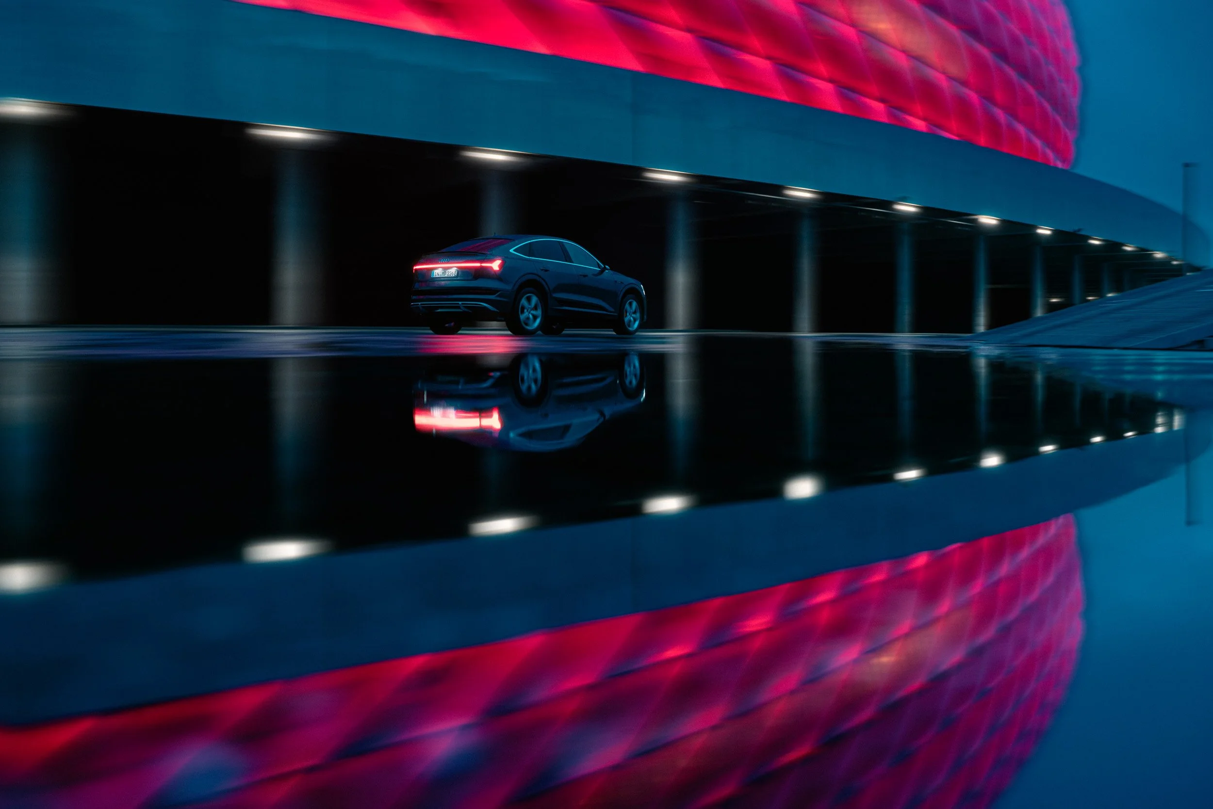 A black luxury car driving on a wet road at night, with a modern building in the background featuring a red and purple illuminated facade, and reflections on a puddle in the foreground.